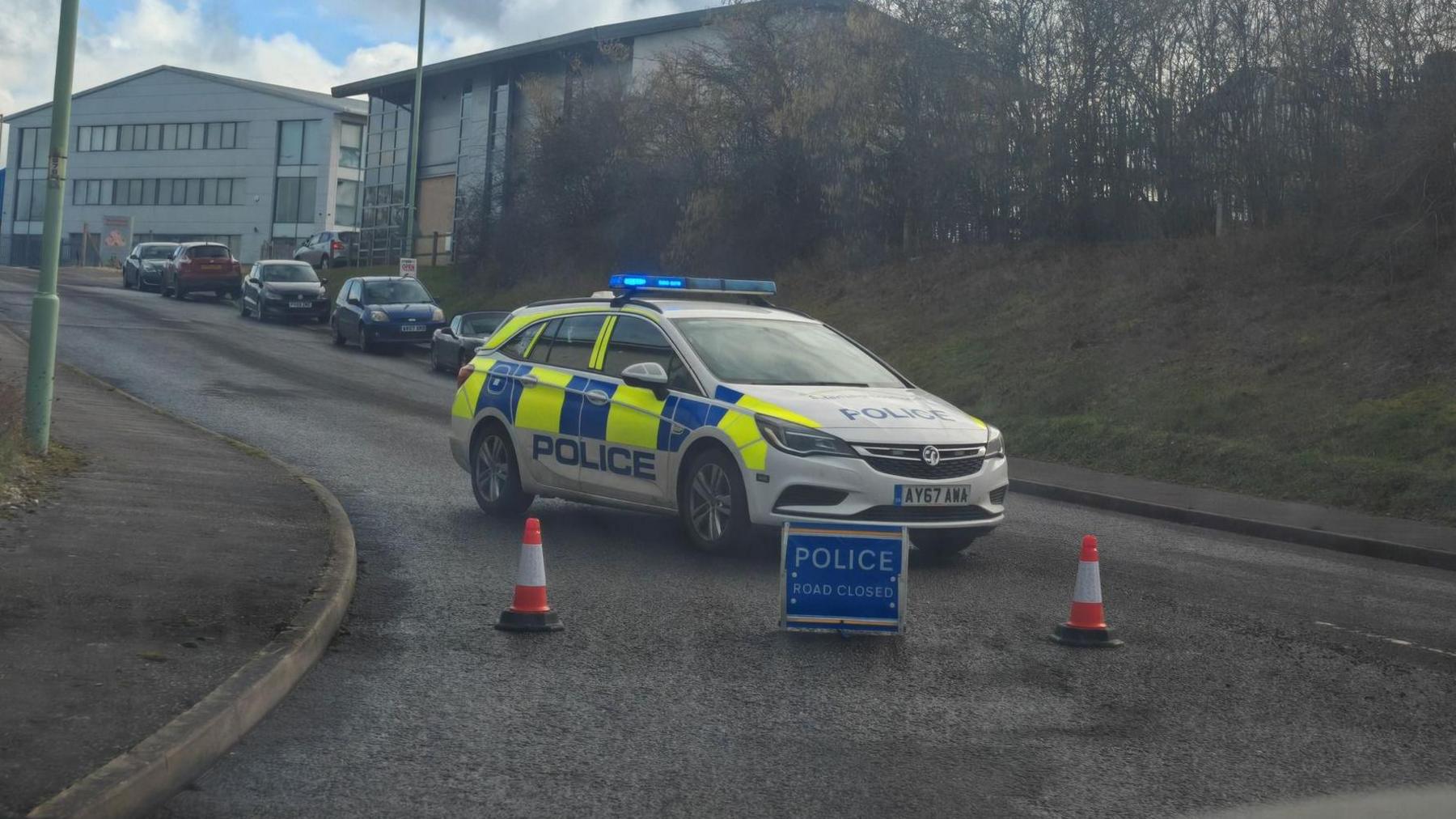 A police car parked in the middle of the road behind a 'police road closed' sign and two traffic cones. Other cars can be seen parked in the background in front of buildings.