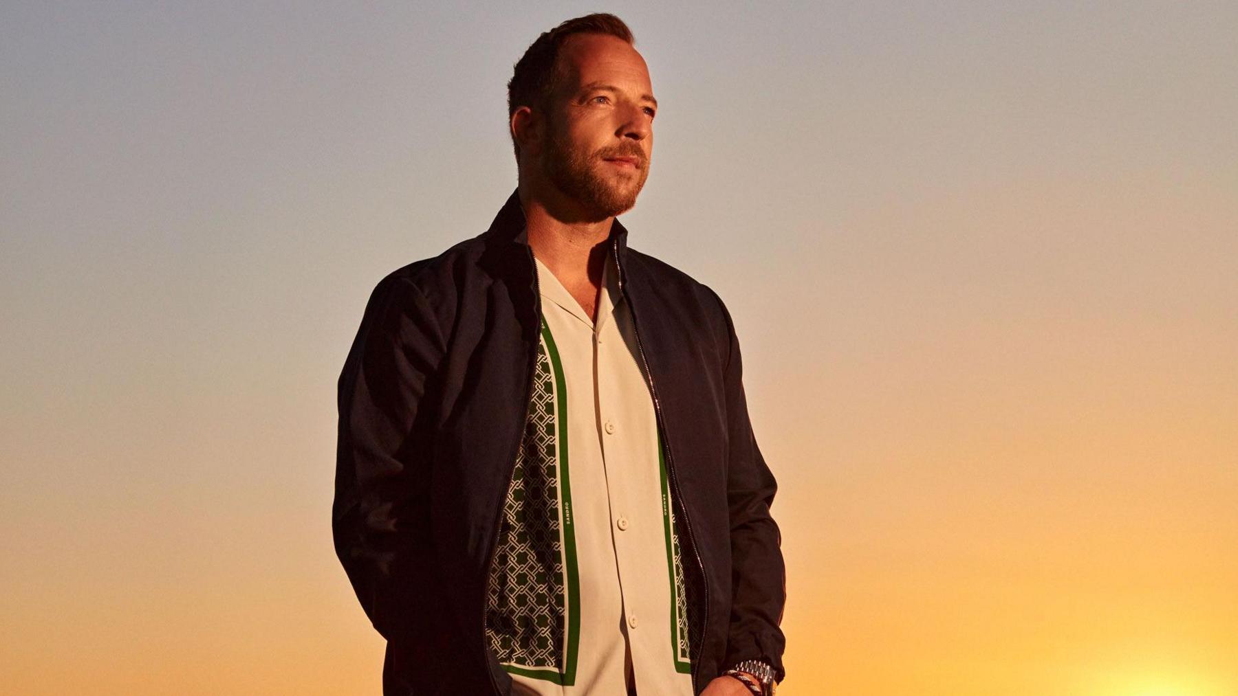 James Morrison, standing, wearing a black jacket, white shirt, with green and black on it, he is looking off to the right, he has a beard and short hair.