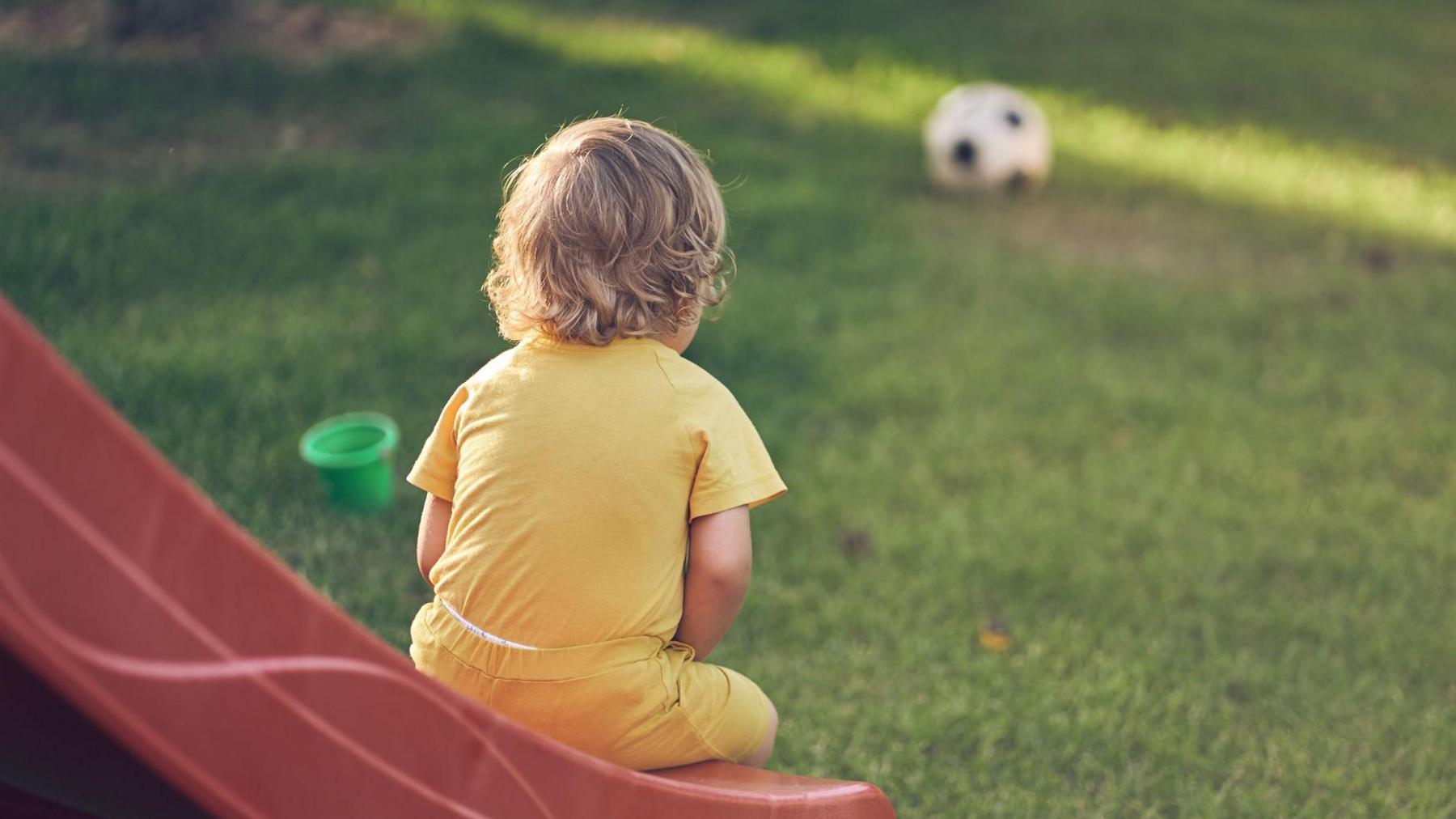 A young boy sits on the end of a slide in a play area, looking at a football