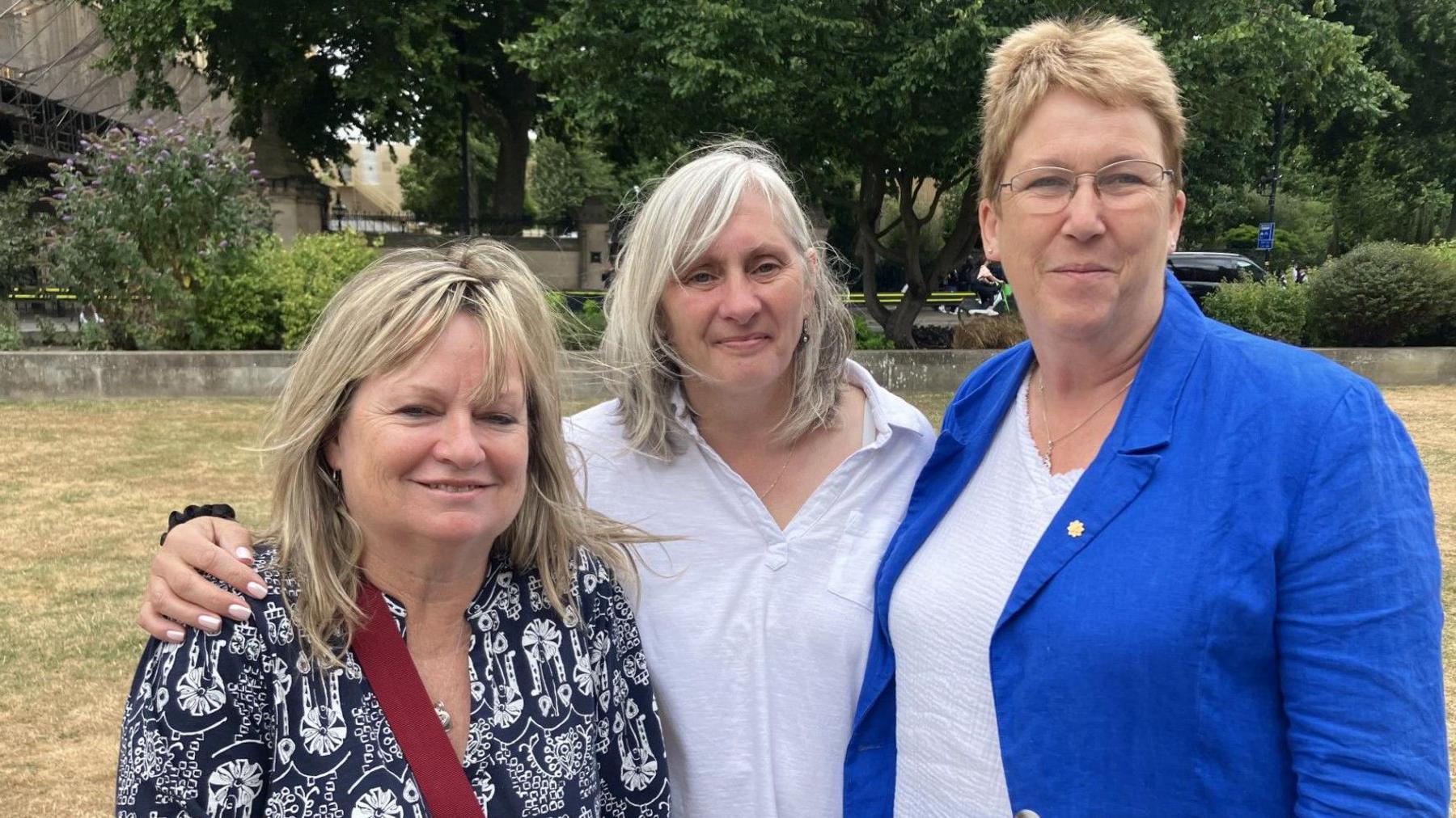 Three women smiling for a photo stood on a patch of grass in a field or a park. The women on the left has blonde hair and is wearing a blue and white top with a red bag strap across her chest. The woman in the middle has grey hair and is wearing a white top, the woman on the right has blonde hair, is wearing glasses and a blue jacket over a white top. 