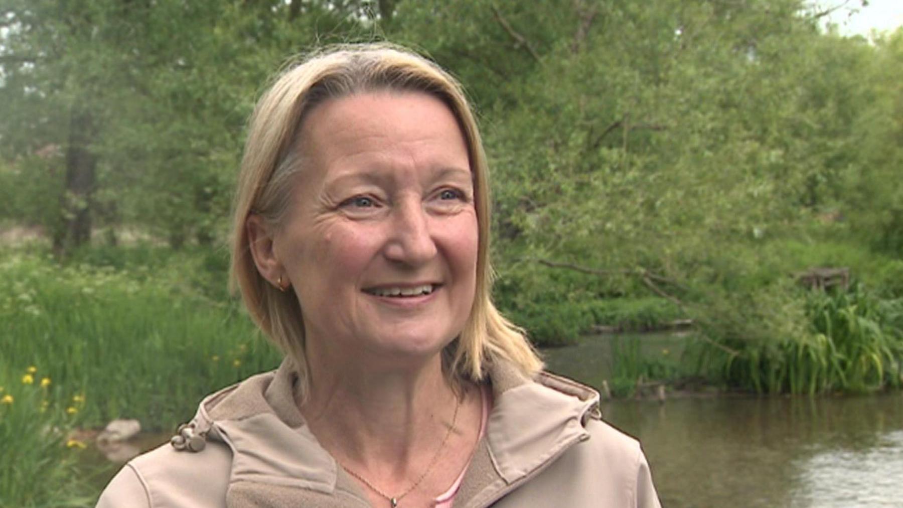 A woman with blond shoulder length hair and a beige coat stands in front of a river, wildflowers and trees are visible in the background.