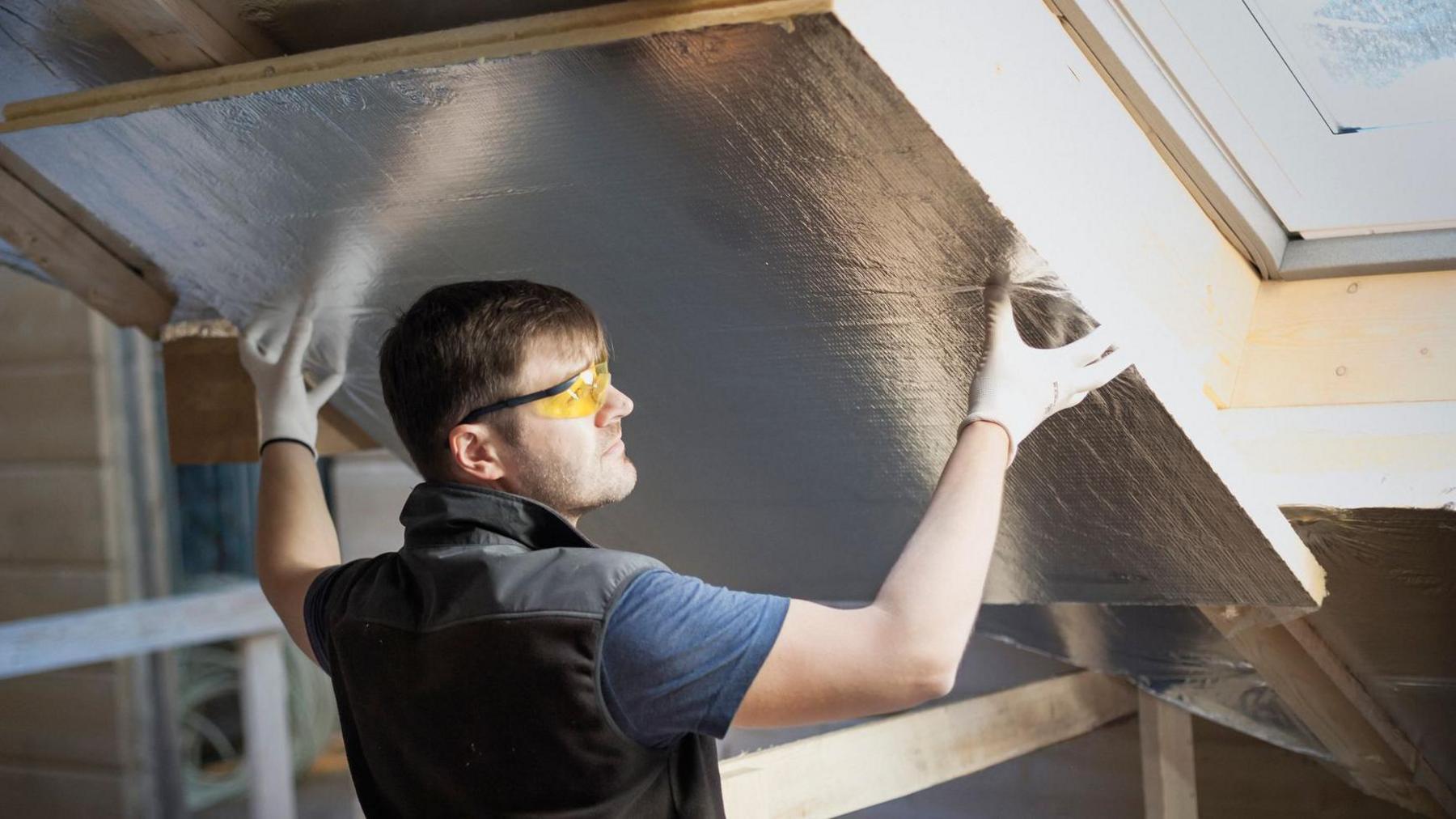 A workman wearing safety specs fits some insulation between roof beams.