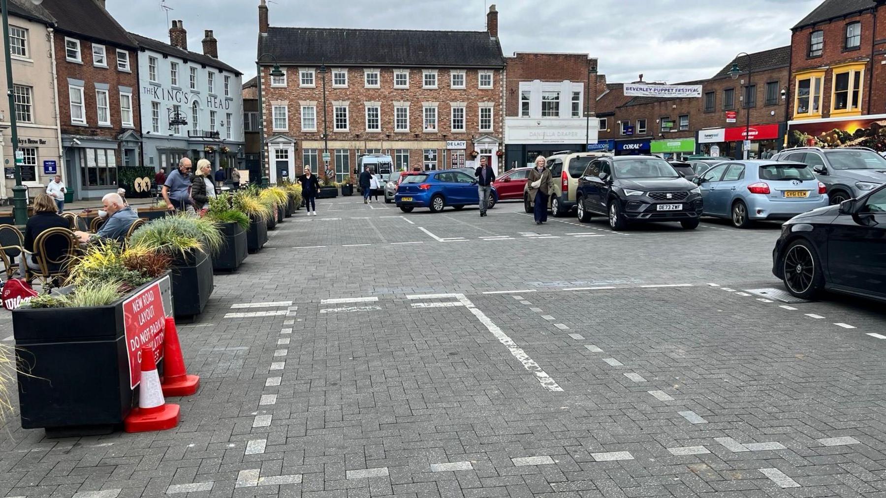 New white road markings in foreground with people, parked cars and historic buildings in background