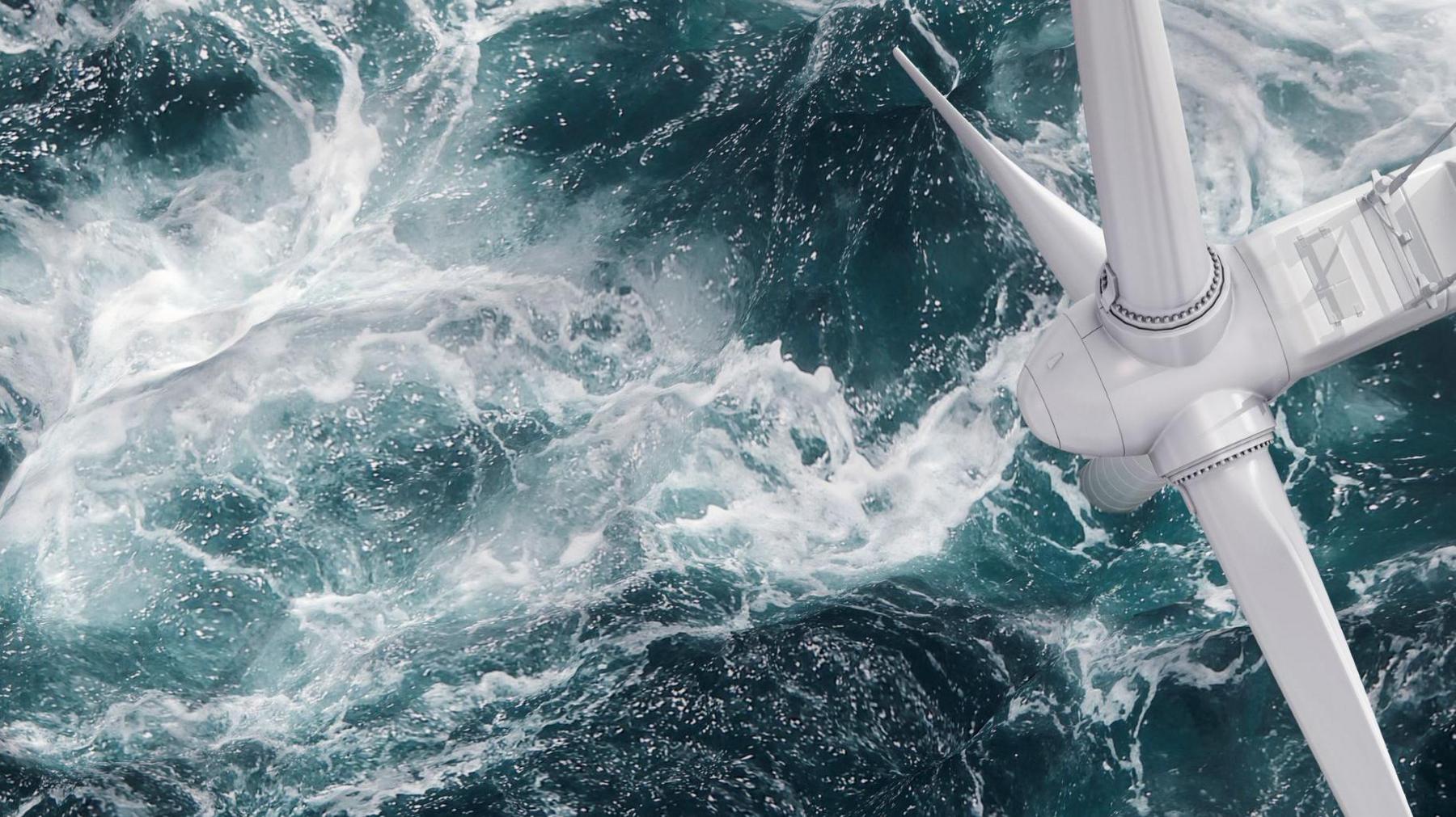 An aerial view of an offshore wind turbine. Parts of the three white blades are visible above the sea, which is a dark aqua green colour. 