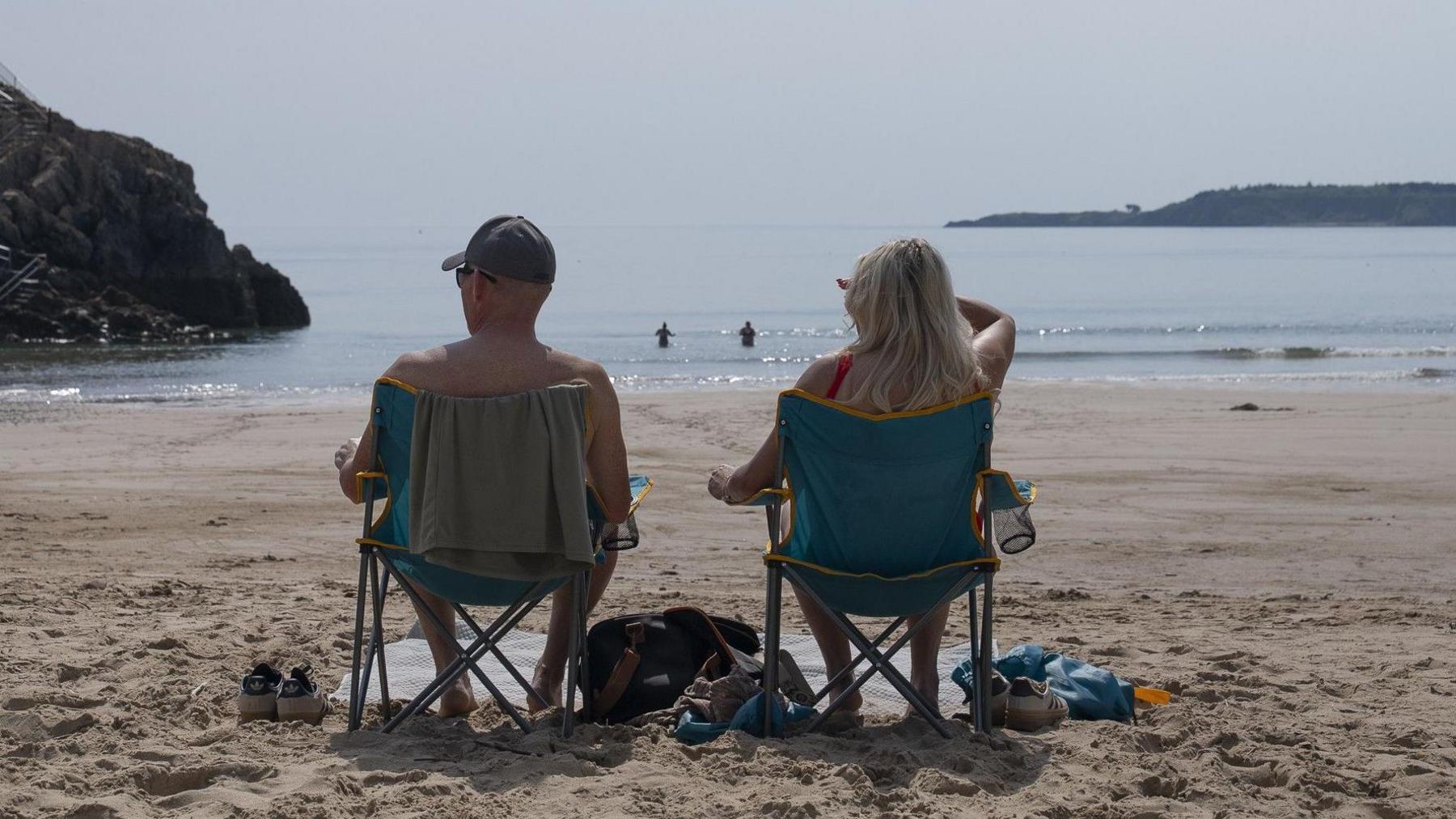 The backs of two people sat on deckchairs. One is a topless man wearing a baseball cap and sunglasses with a towel on the back of his blue chair. The other is a woman with long blonde hair in a red swimsuit, sat in a matching blue chair with one hand up to shield her eyes from the sun. The beach is sandy, and they have bags and shoes beside them and are looking out at the sea where two more people are swimming. There are cliffs on either side.