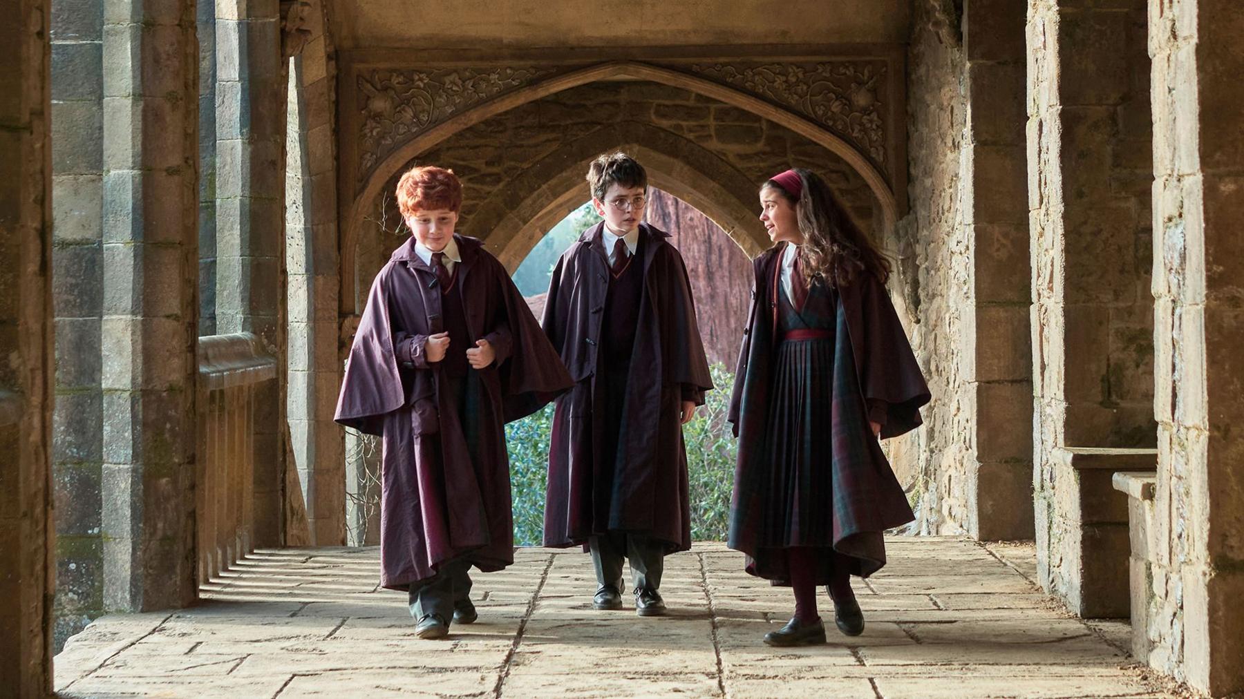 Alastair Stout as Ron Weasley, Dominic McLaughlin as Harry Potter and Arabella Stanton as Hermione Granger in the HBO YV series walking along a cloister of fictional Hogwarts school set.