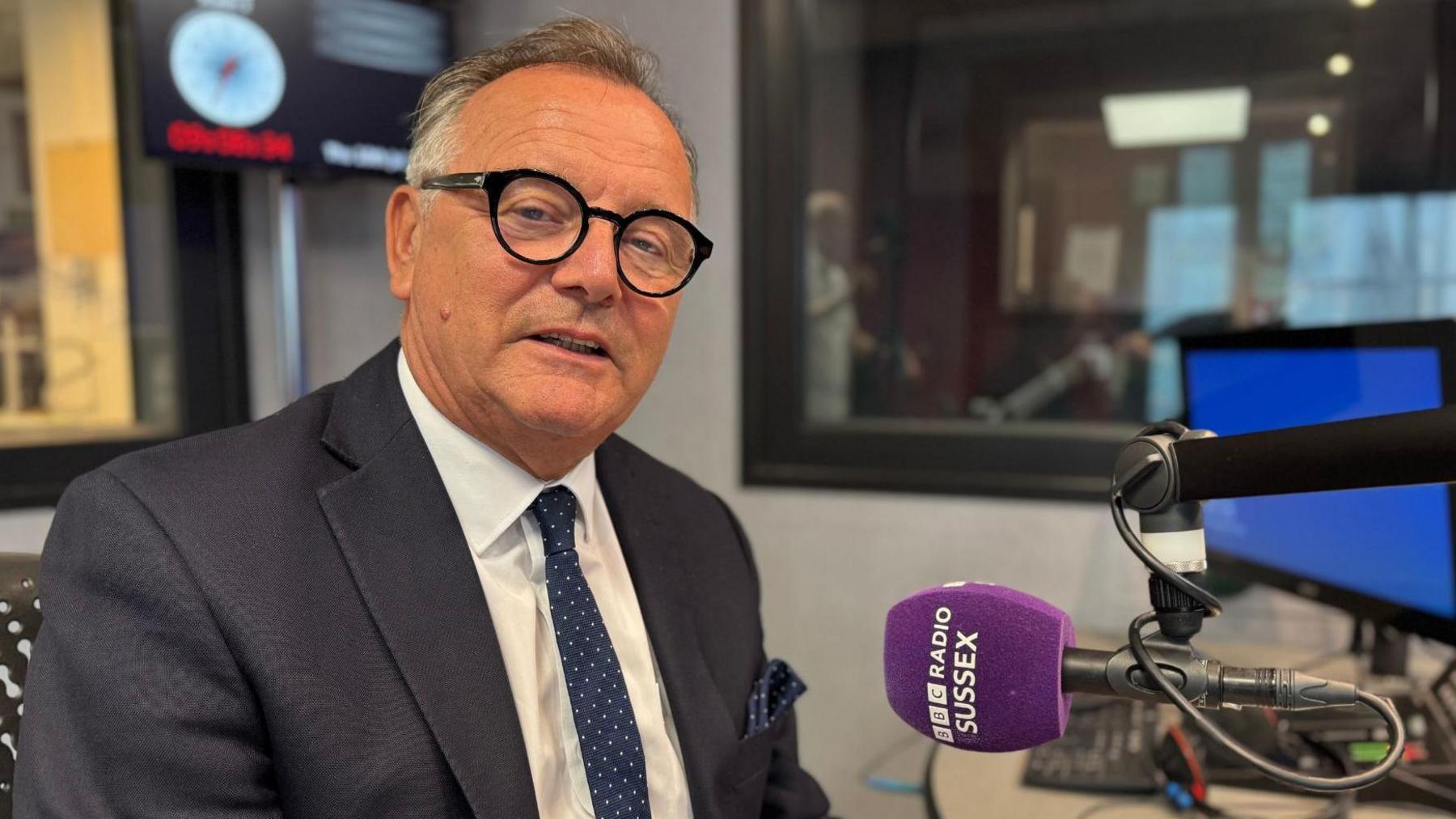 A man wearing a grey suit jacket, white shirt and dark blue tie with white spots on. He is sat in a chair in a radio studio looking into the camera. In front of him is a purple microphone with the words BBC RADIO SUSSEX written on it in white lettering