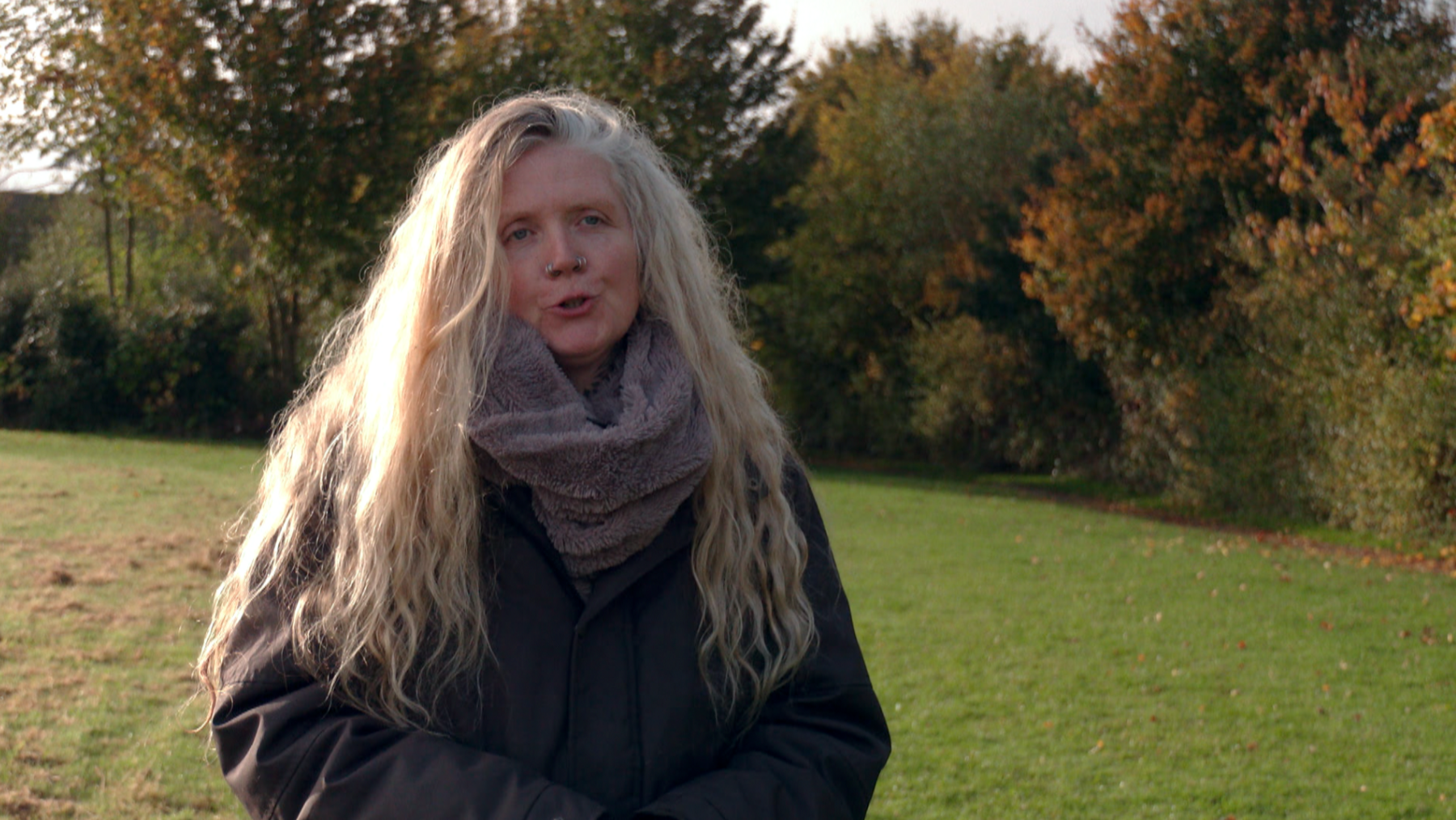 Campaigner Cathy Scott, wearing a grey coat and light grey scarf, is stood in a field with trees in the background. She has long hair and nose piercings on both nostrils.