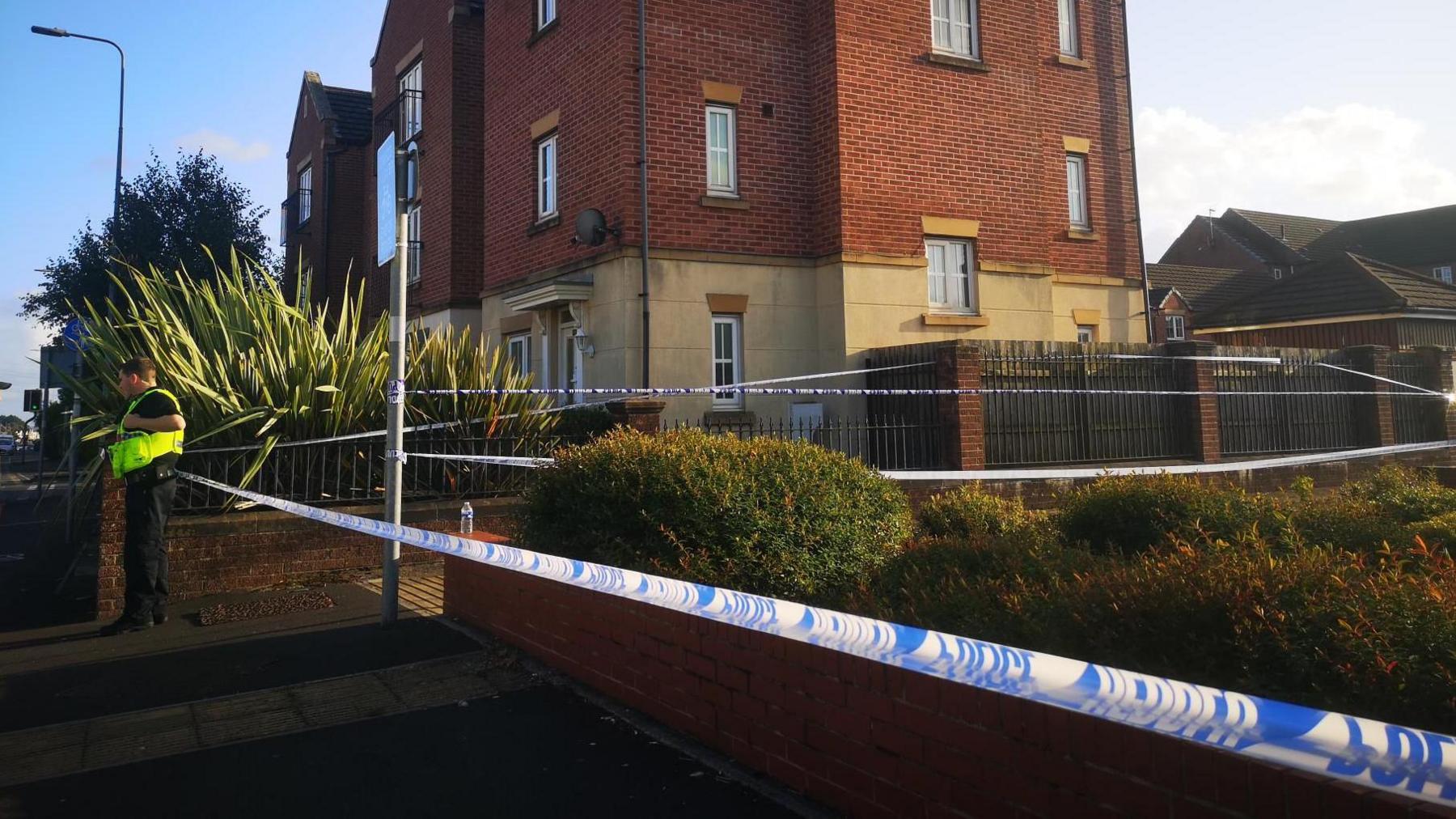 Police tape zigzags up an entrance way to a building, visible between the plants and shrubs. A police officer stands on the left with their high-vis yellow jacket on. 