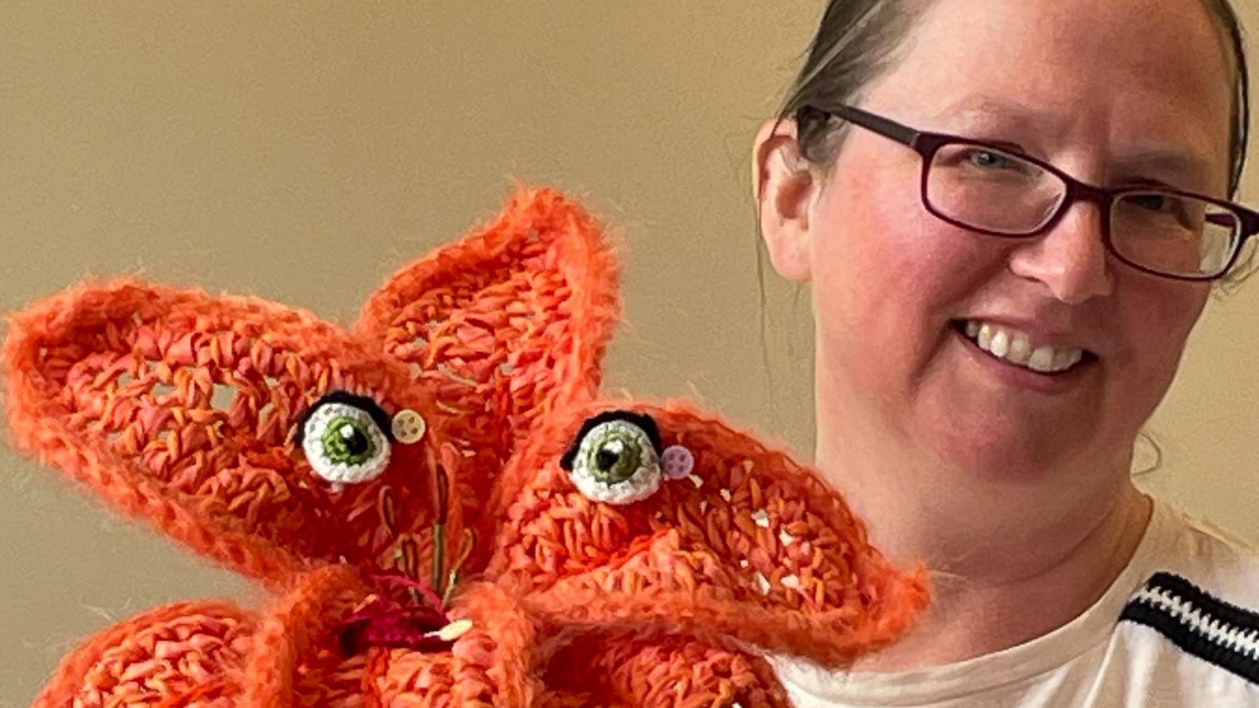 A woman wearing glasses with her hair tied back smiles at the camera. Next to her is an orange knitted flower which has crafted eyes.