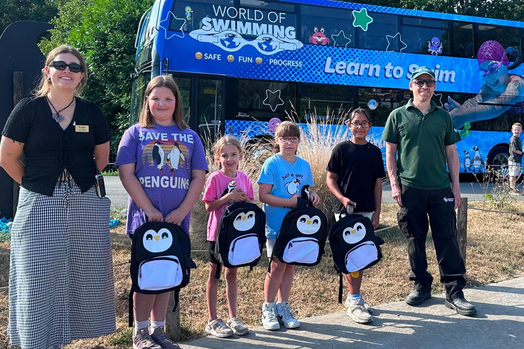 Children and staff pose with penguin backpacks in front of the World of Swimming bus.