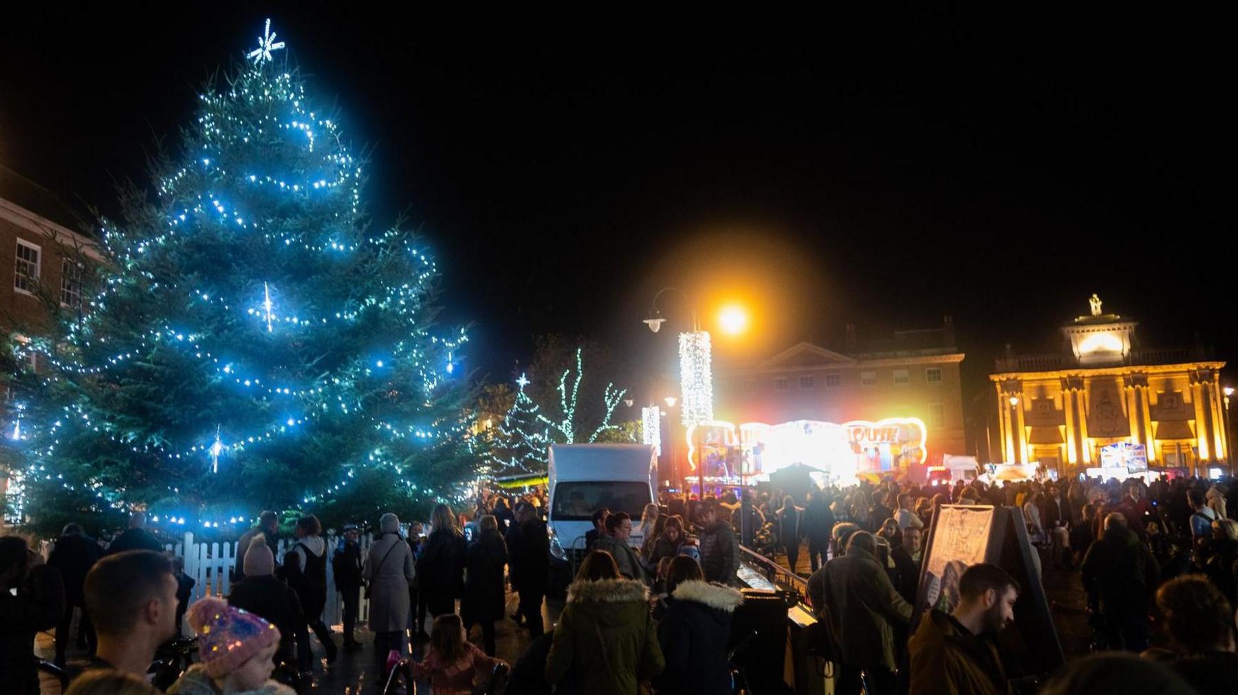 A night time Christmas light switch-on in King's Lynn, showing a light Christmas tree, light up buildings, people standing bout, a white van, and street lights.