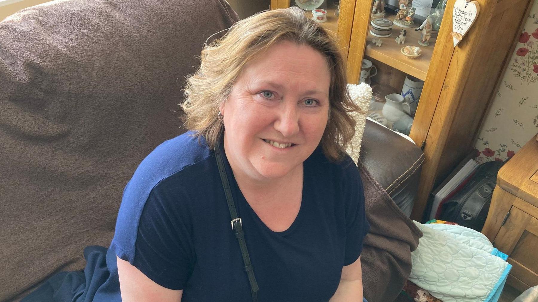 A woman looks up at the camera while sitting on a brown leather sofa, next to a cabinet. She is wearing a blue T-shirt and has chin-length reddish-brown hair. She has a handbag strap over her shoulder and is smiling.