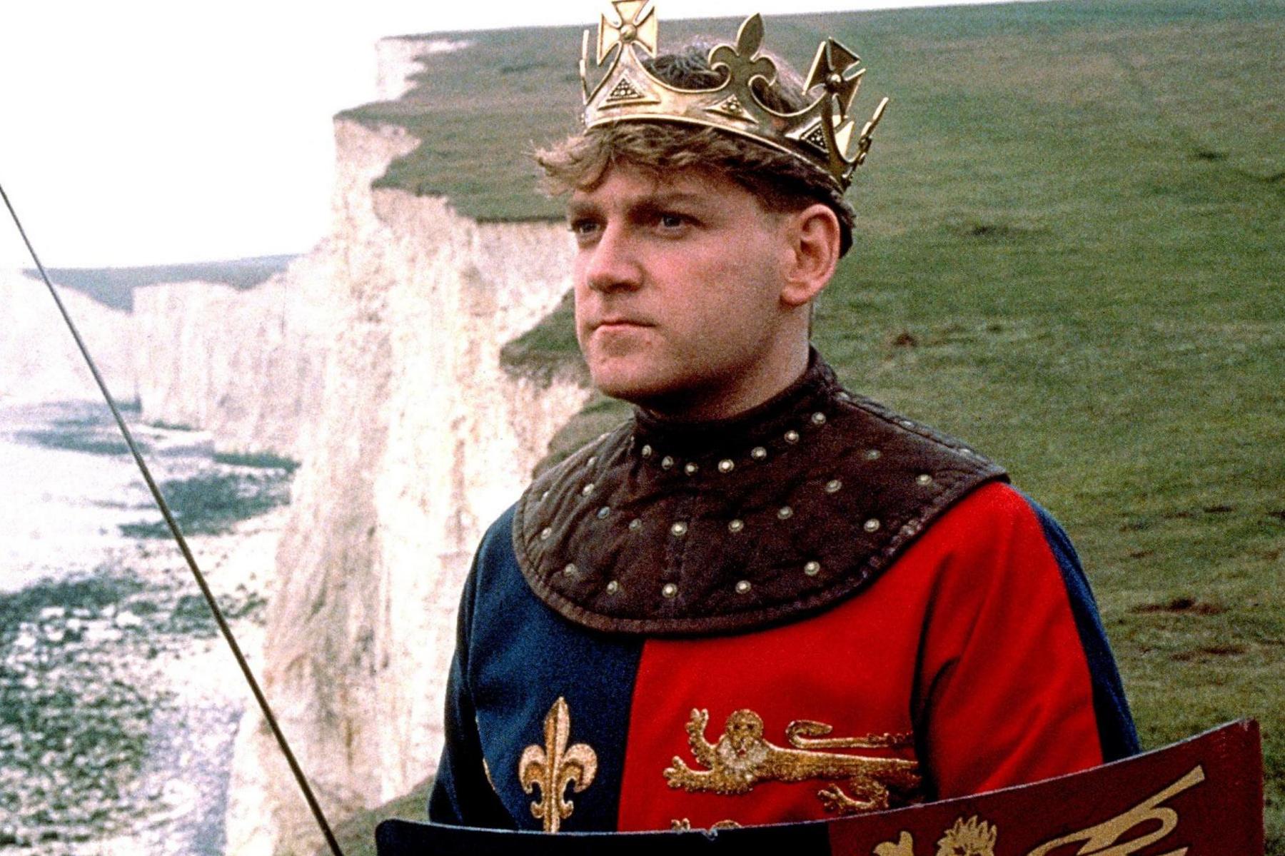 Kenneth Branagh with gold crown, dressed in blue and red regalia, with sword and shield, standing a cliff on  edge of coast.