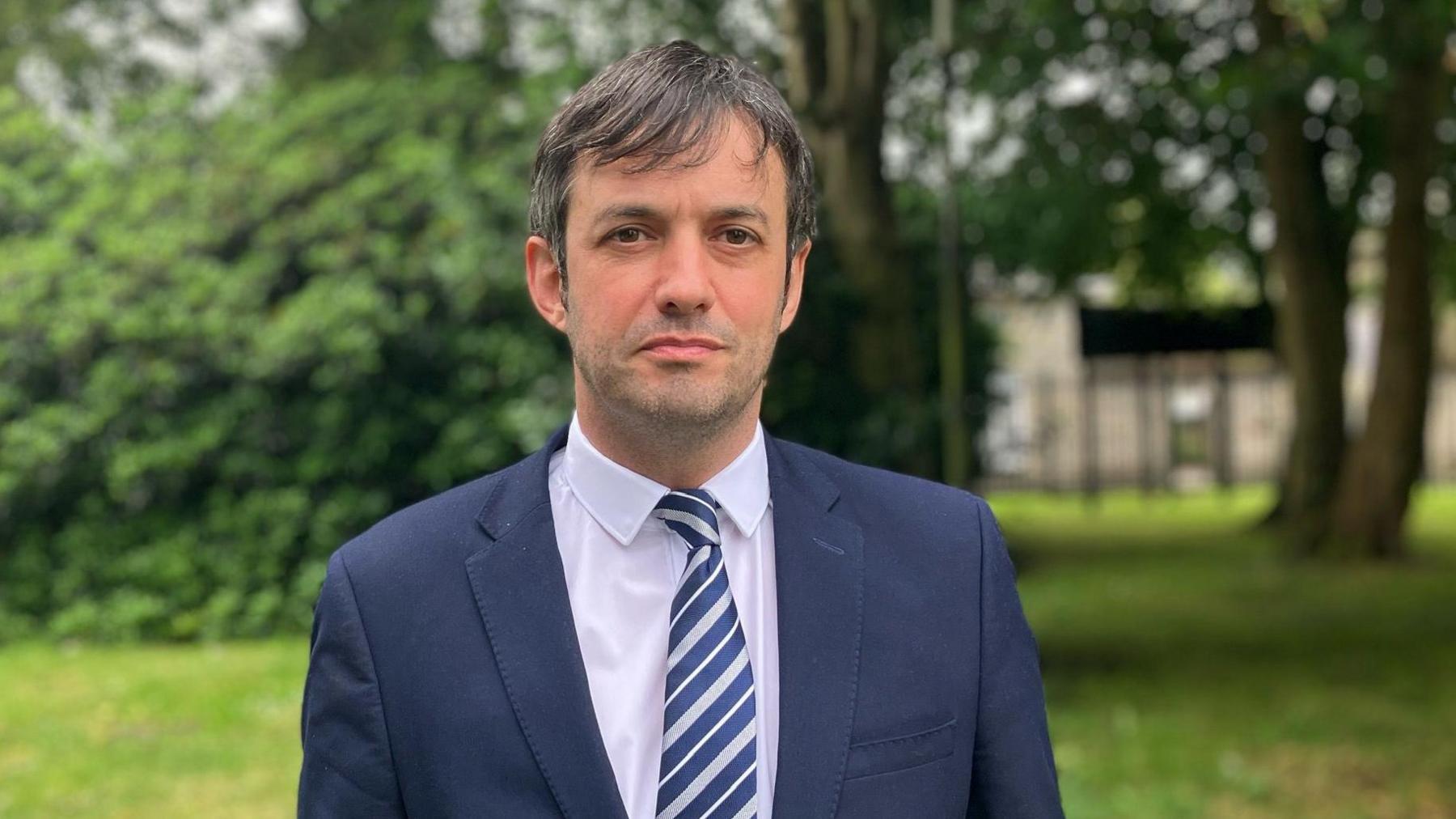 Man in navy jacket with white shirt and blue and white striped tie standing in front of trees