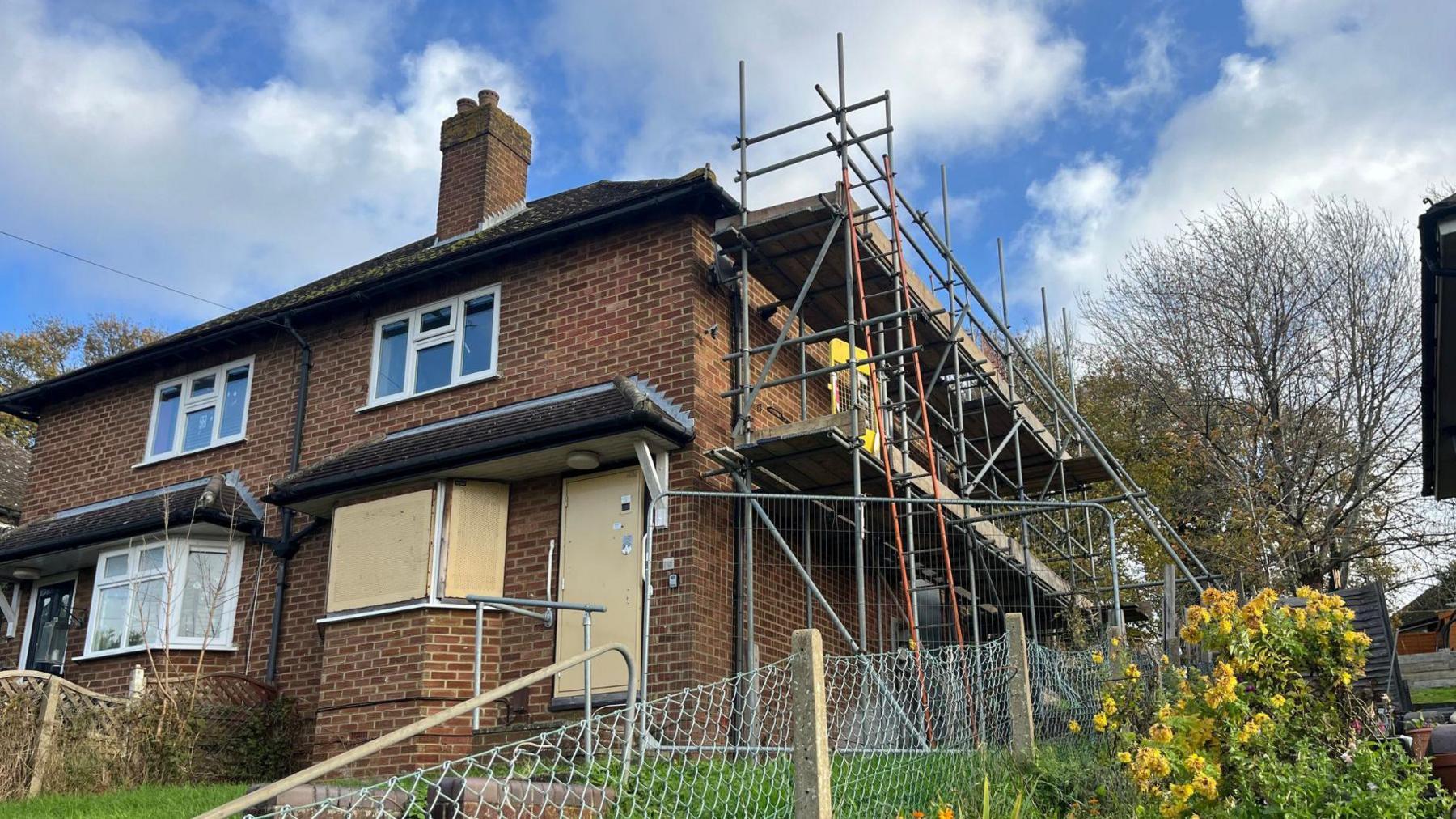 A semi-detached terraced house with boarded up ground floor windows and scaffolding on its right wall