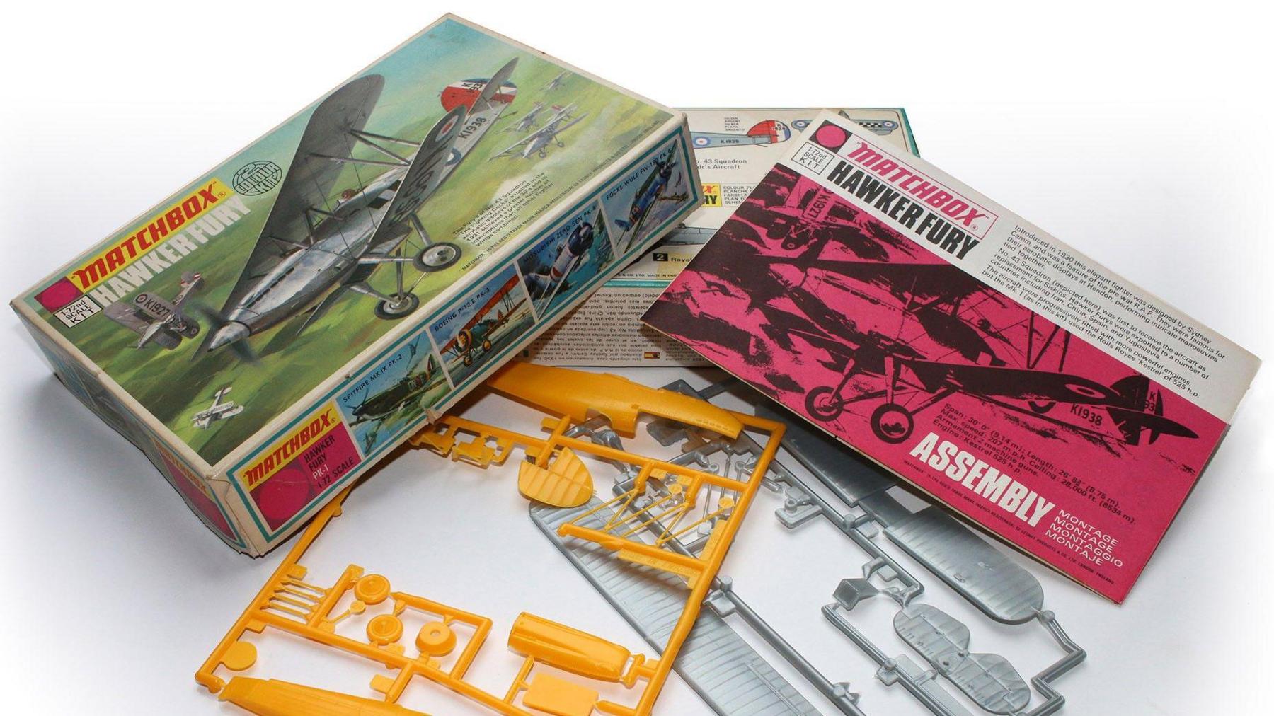 A multi-coloured Matchbox model box adorned with a painting of a Hawker Fury lying on a white table.  A pink and white assembly instruction booklet lies to the right of the box. Lying in front of the box are yellow and grey parts of the model kit.