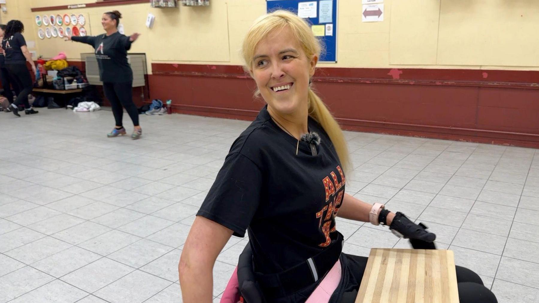 Claire Booth is in a dance studio with other dancers in the background. She has long blonde hair and is wearing a black t-shirt and black gloves with a wooden board on her knee