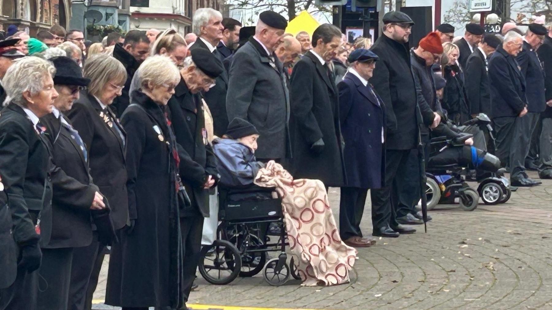 A crowd of people wearing dark colours stood at a Remembrance service in   Ilkeston, Derbyshire. In the middle of them is Donald Rose, aged 109, who is in a wheelchair with a blanket over his legs.