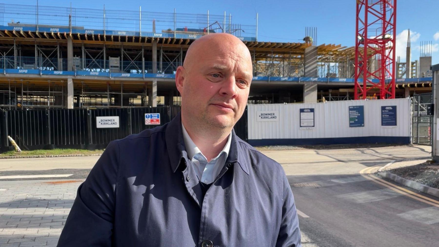 A man wearing blue coat standing in front of a construction site, which is cordoned off, with the bottom of a red crane behind fencing and concrete columns