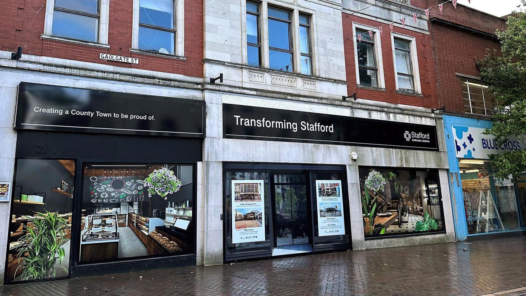 A shop unit with white stone and black windows and boards. On the boards over the windows, there are photos of what the site will eventually look like. This includes a photo of a bakery and plants with a large staircase. A sign above the windows reads "Transforming Stafford"