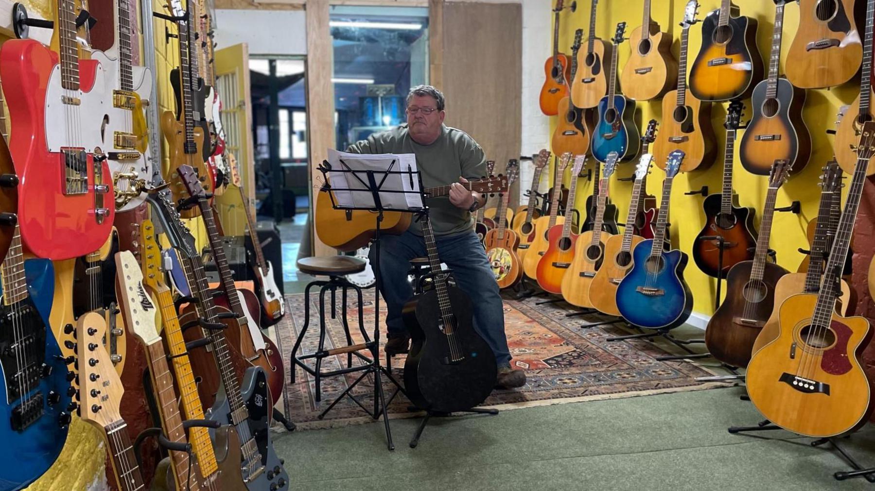 A heavy set man sits holding a guitar ready to play in a music shop. Acoustic and electric guitars are lined up on both sides of the shop