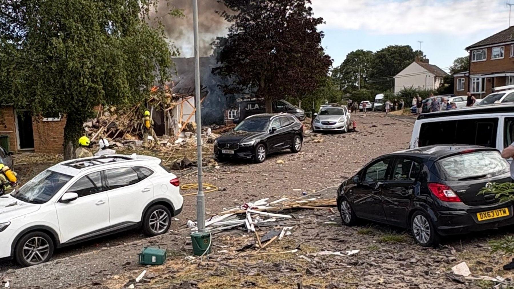 Firecrews attending a fire at a house which has been completely destroyed. Car have their roofs caved in and there is debris all over the street.