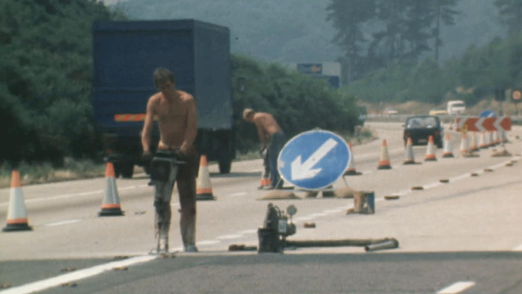 Two bare-chested men in jeans drill into the motorway in the 1970s. Traffic cones are laid out, a lorry is parked in the background. A blue Austin Metro is driving in the distance.