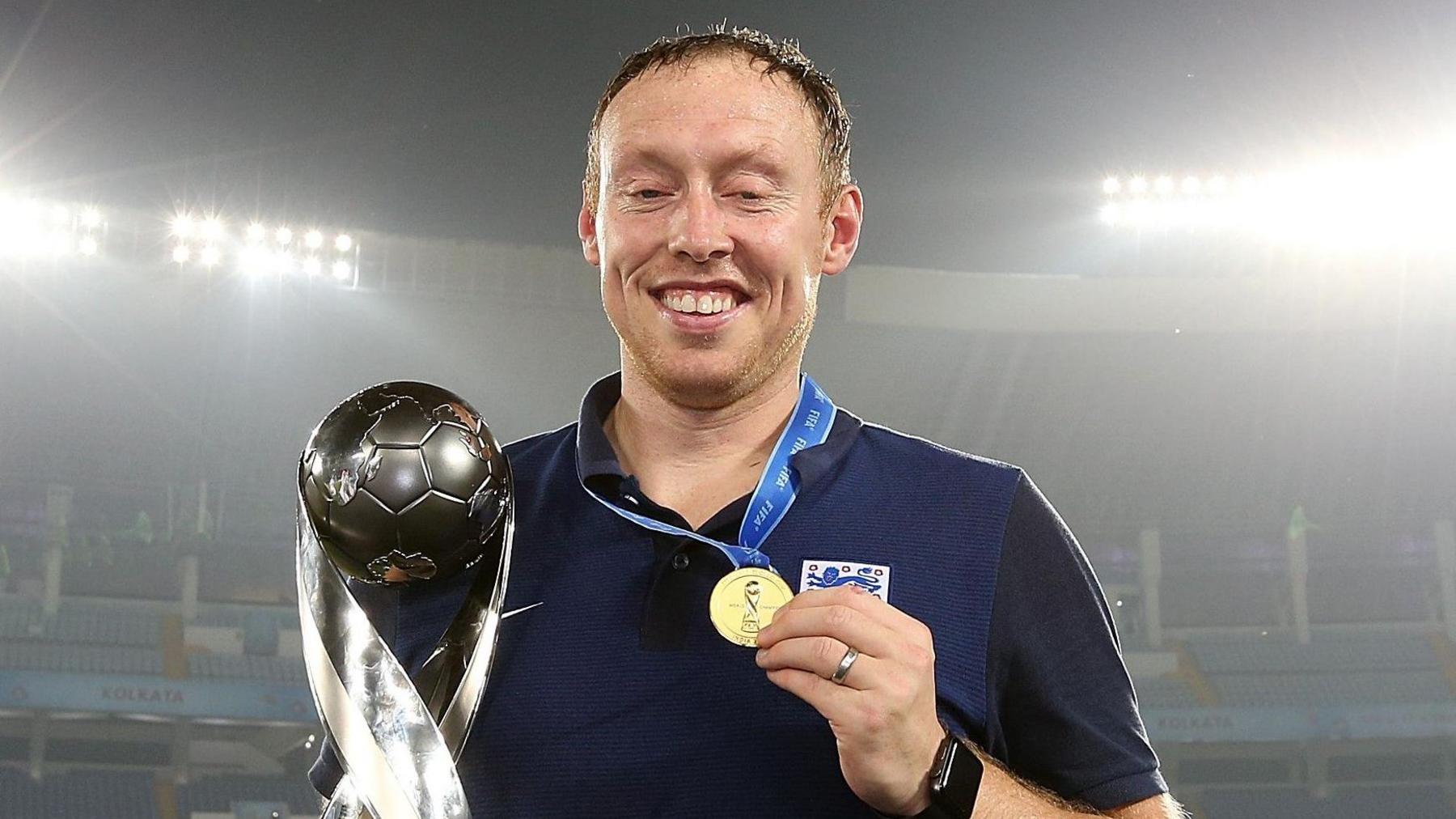 Steve Cooper holds the silver Under-17 trophy in his right hand and raises his gold medal in his left hand