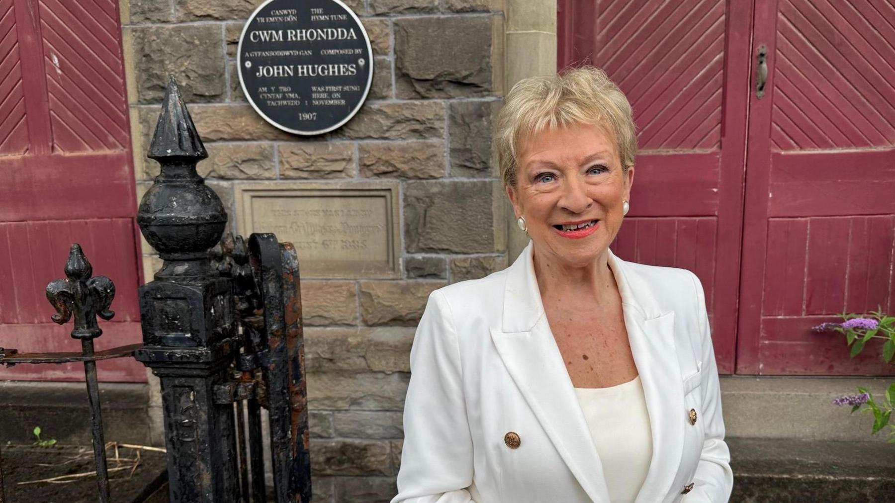 Beverley Humphreys is standing in front of the chapel and smiling to the camera. She has short, light blonde hair and a white jacket with gold buttons and a white medium cut blouse underneath.