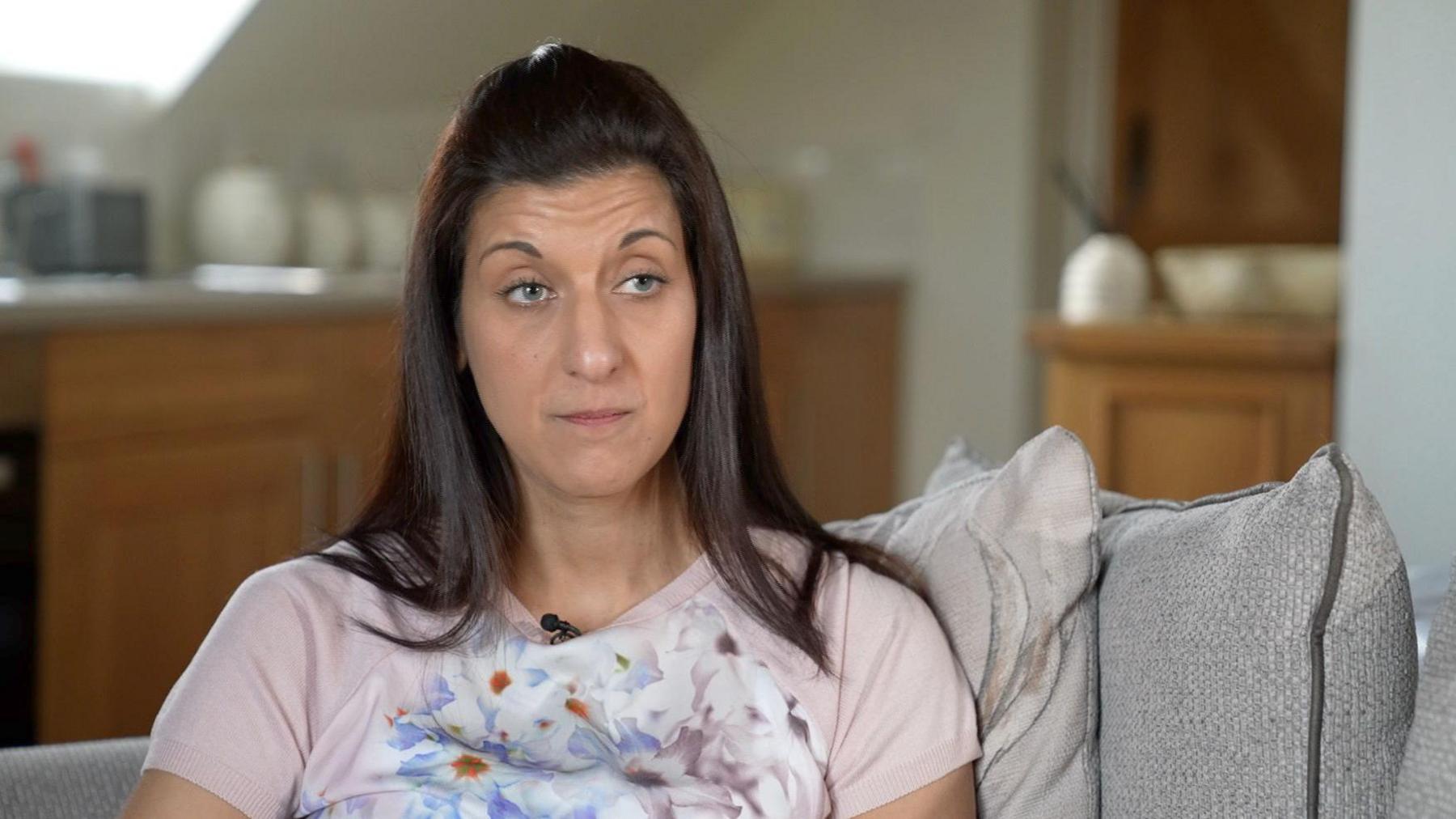 Rebecca Kalam sitting on a sofa with a kitchen in the background. She is wearing a pink T-shift with a flowery pattern and has long, black hair, tied back in a clip.