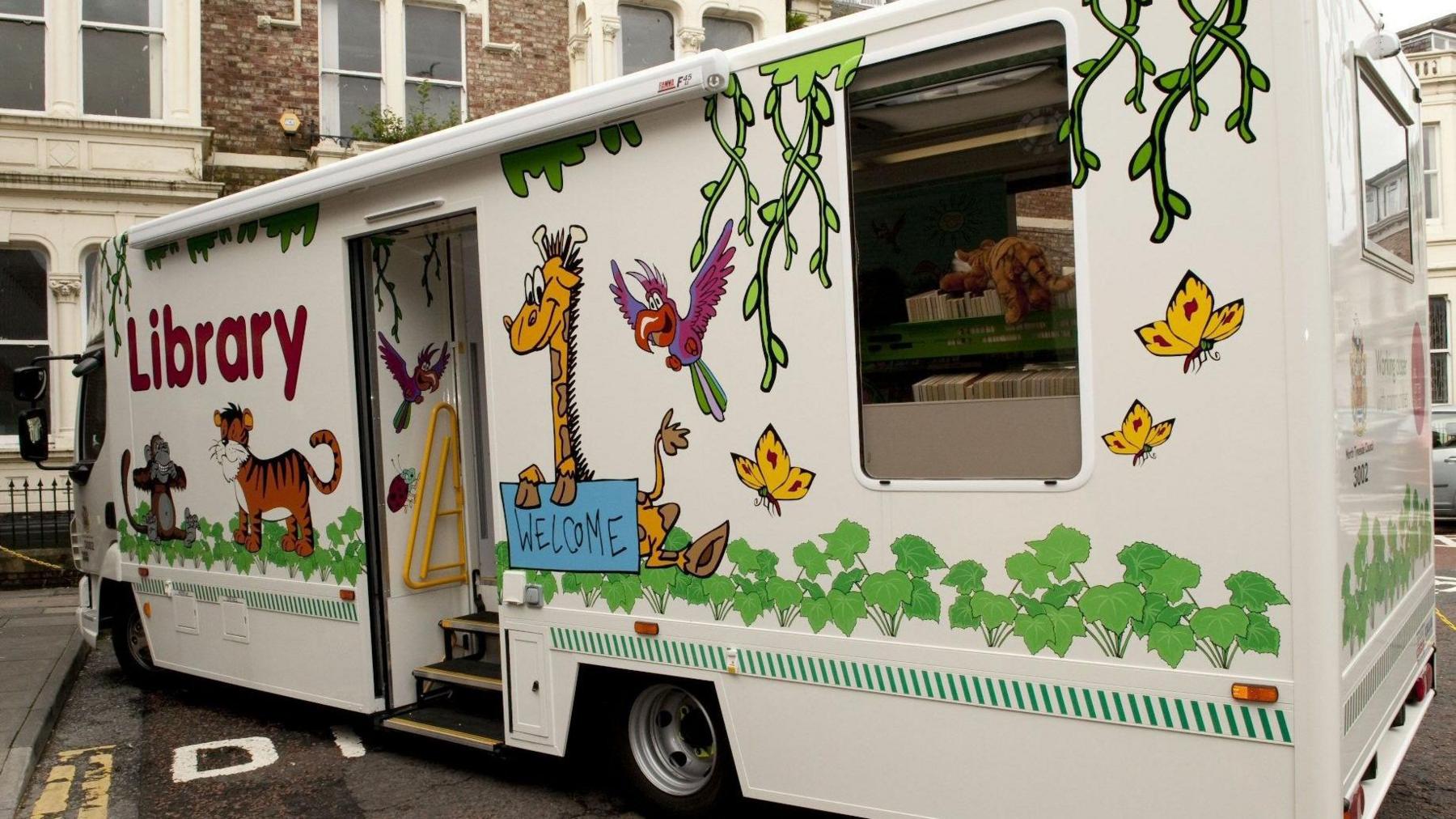 A campervan with animal characters painted on the side, together with a sign saying "library" and one saying "welcome". Through the side window, books can be seen ordered on shelves inside, with some stuffed toys.