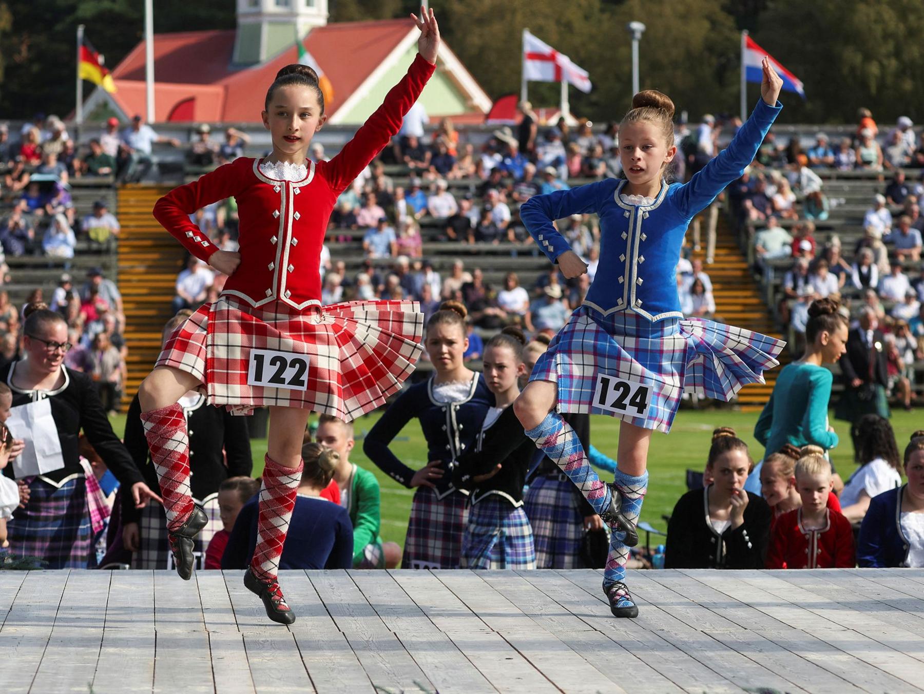 Girls dancing in traditional outfits, one red and one blue, with spectators behind them 