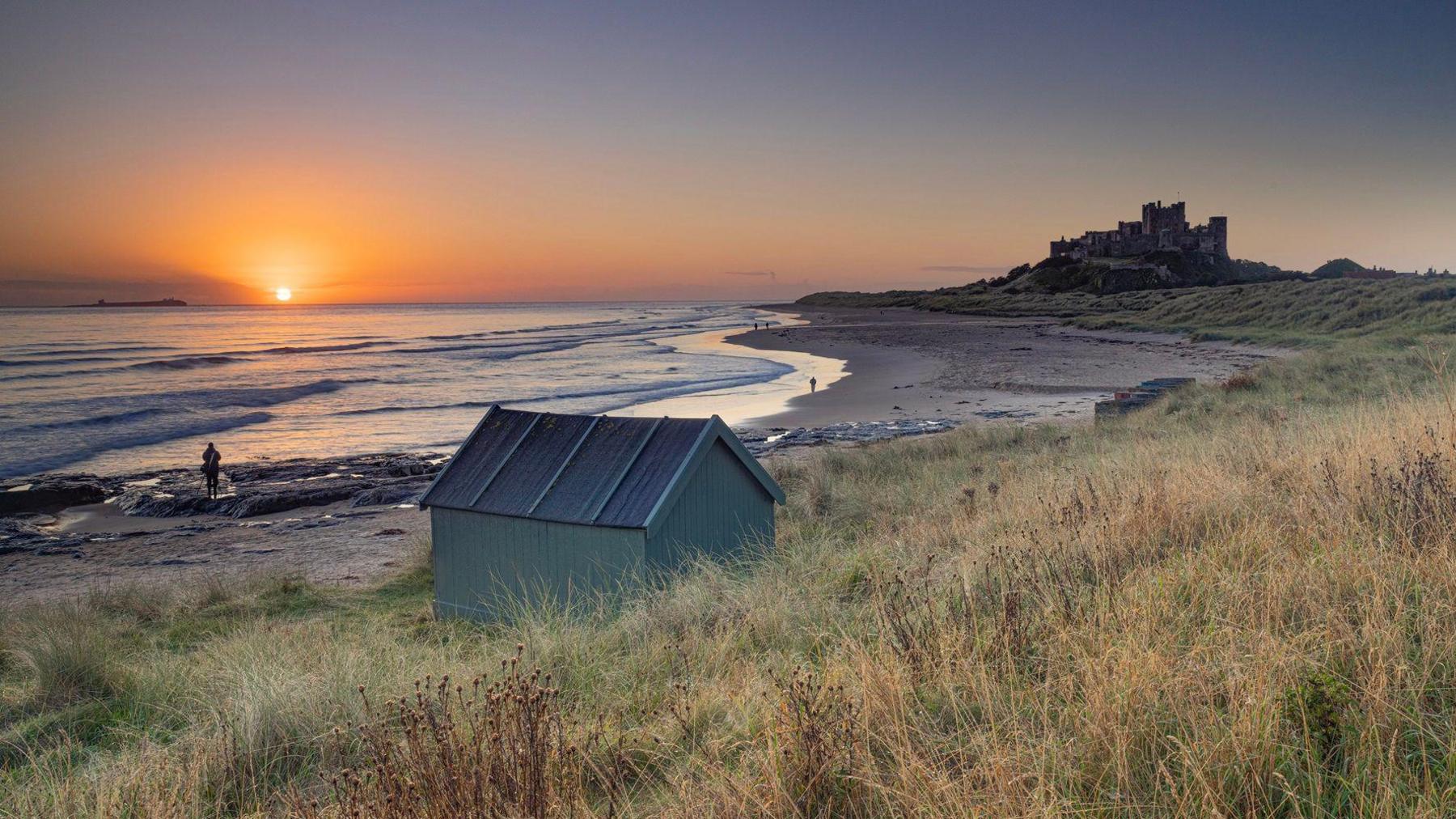 The sun rising over the sea. The sky is orange around the sun, transitioning to a darker blue higher up. A green beach hut is in the foreground, with the beach and sea in front of it. The silhouette of a person can be seen standing on a rock on the beach. The outline of Bamburgh Castle is the to right of the photo.