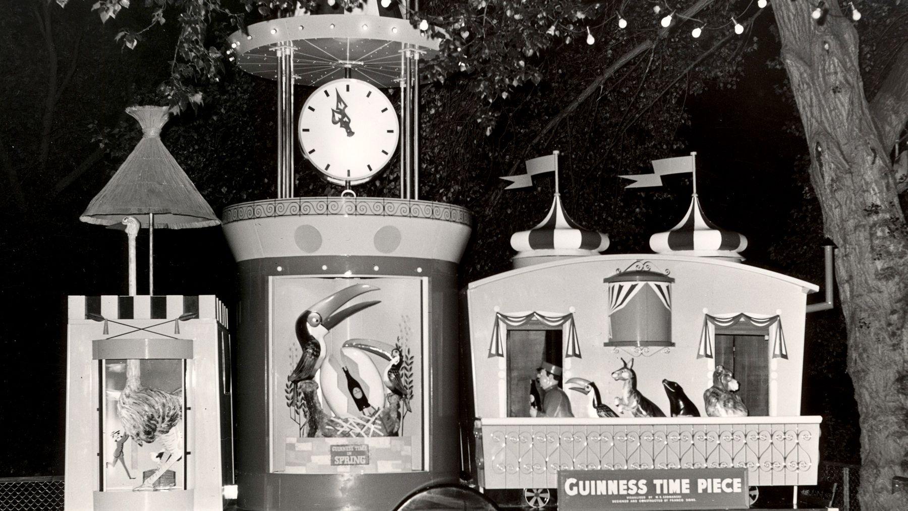 A black and white image of the guinness clock in the 1950s at the light festival. The vertical timepiece has a clock on top with painted toucans below. Next to it is a model caravan with flags on top and a model zoo keeper leading animals thorugh a door.