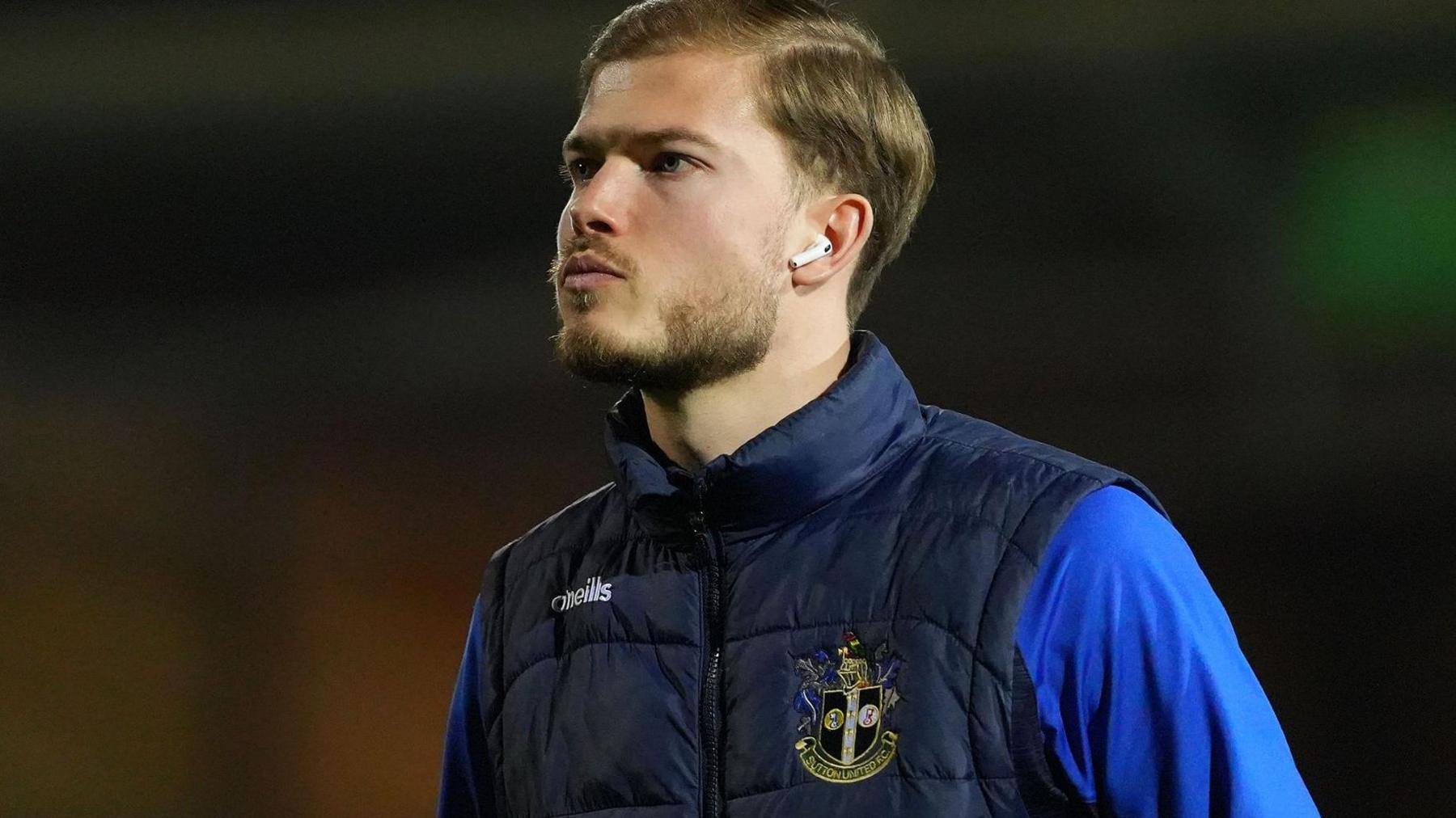Lewis Simper wearing ear buds and a navy blue gilet with the Sutton United crest on it