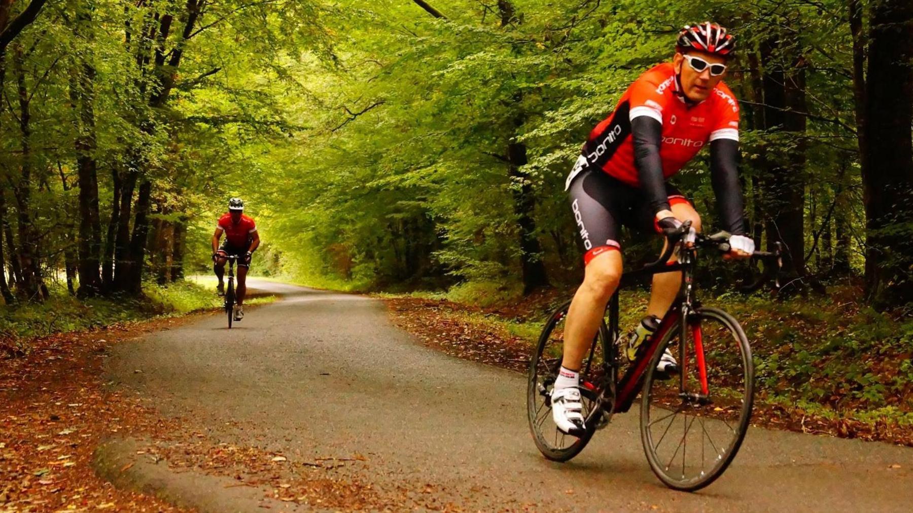 Two cyclists, on a road, with leaves, and trees, riding along the road. They are wearing red and black tops and bottoms. The man in the front has glasses on and a helmet. 