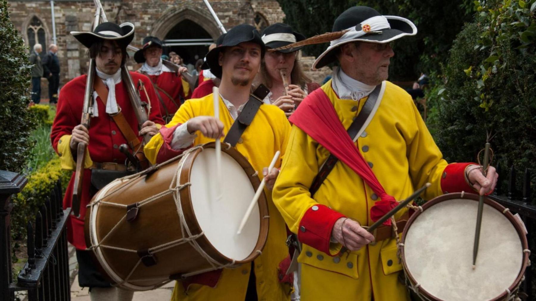 A procession of Wimborne Militia in period costume walking towards the camera. The two leading members are dressed in yellow coats and black hats. Each is playing a large drum hanging over their shoulders.