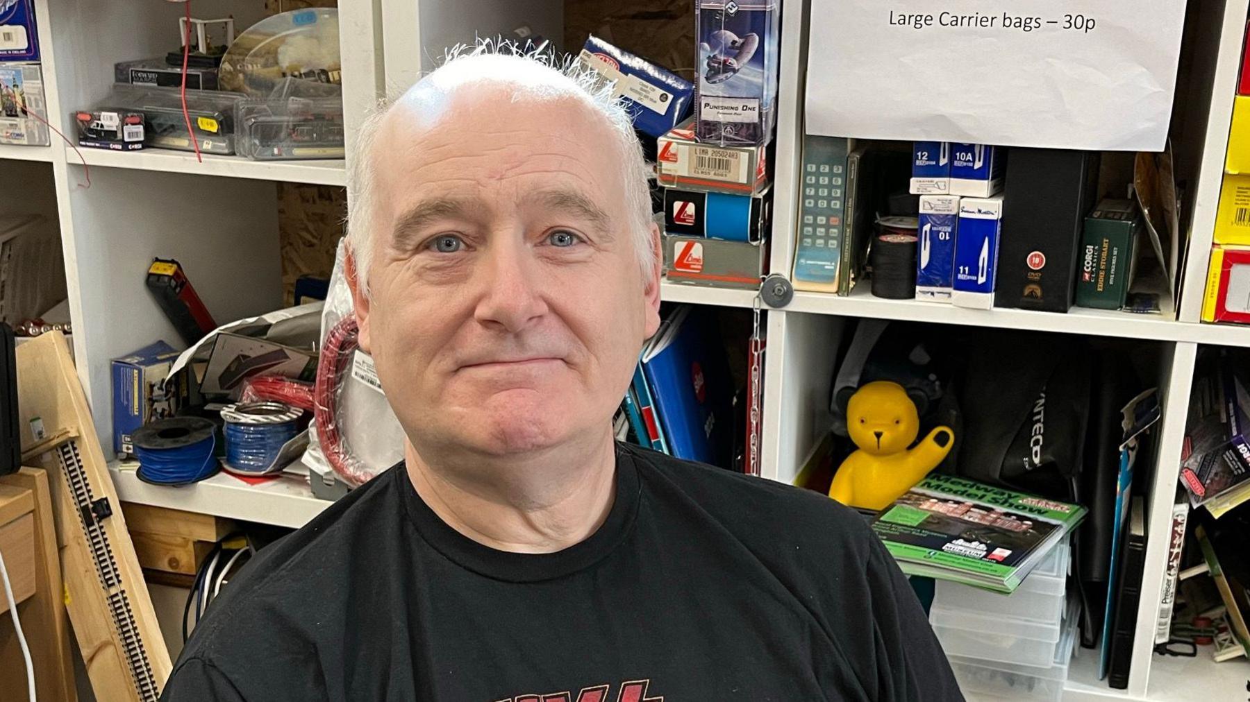 A man with short white hair smiles at the camera. He is wearing a black t-shirt. Behind him are white storage containers filled with bric-a-brac.