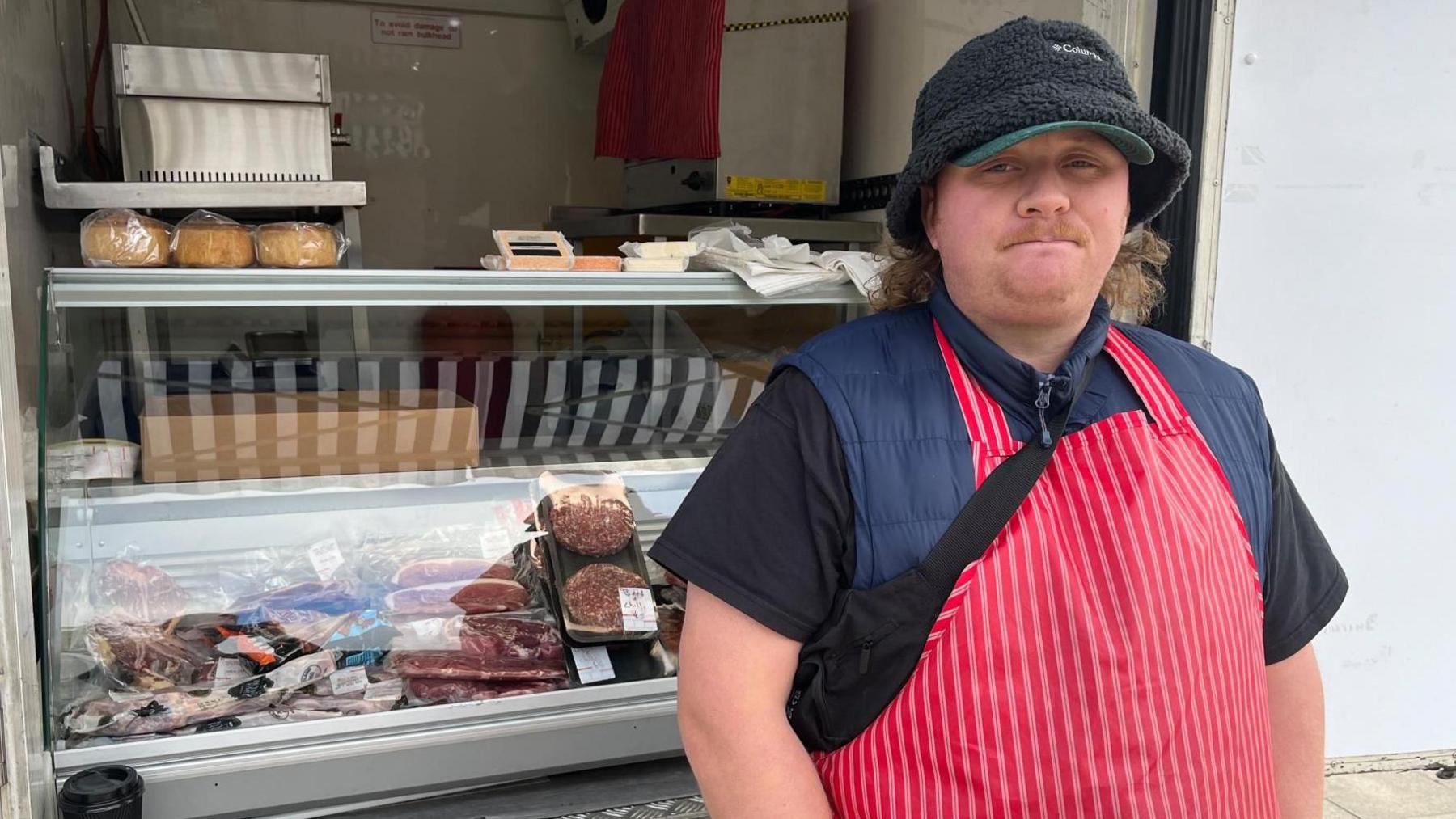 A man wearing two hats, a body warmer and a red and white butcher's apron. He is standing in front of a meat chiller cabinet which is loaded with stakes and burgers.