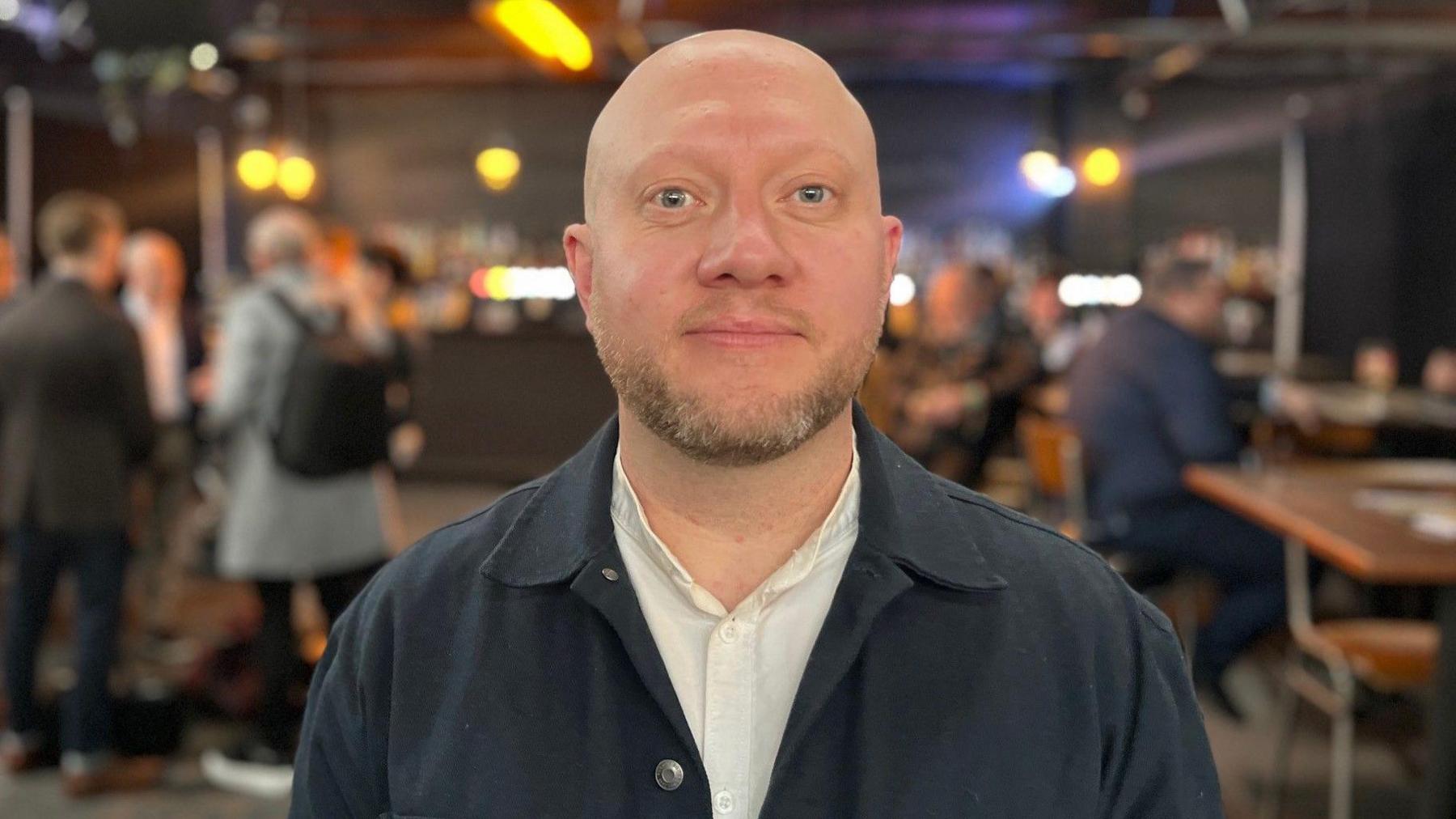 A shaven-headed man with a short beard is wearing a black jacket and pale-coloured shirt, and is standing in a bar.