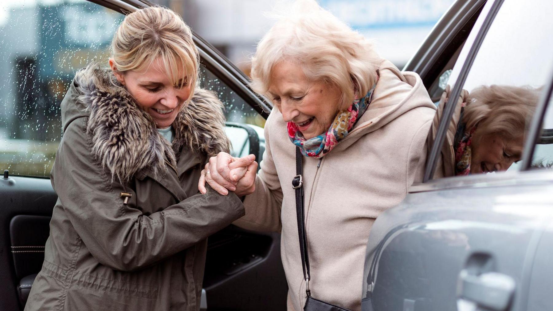 A blonde woman in a furry parka helps a grey haired senior woman in a cream top and scarf out of a car as she takes her to the shops