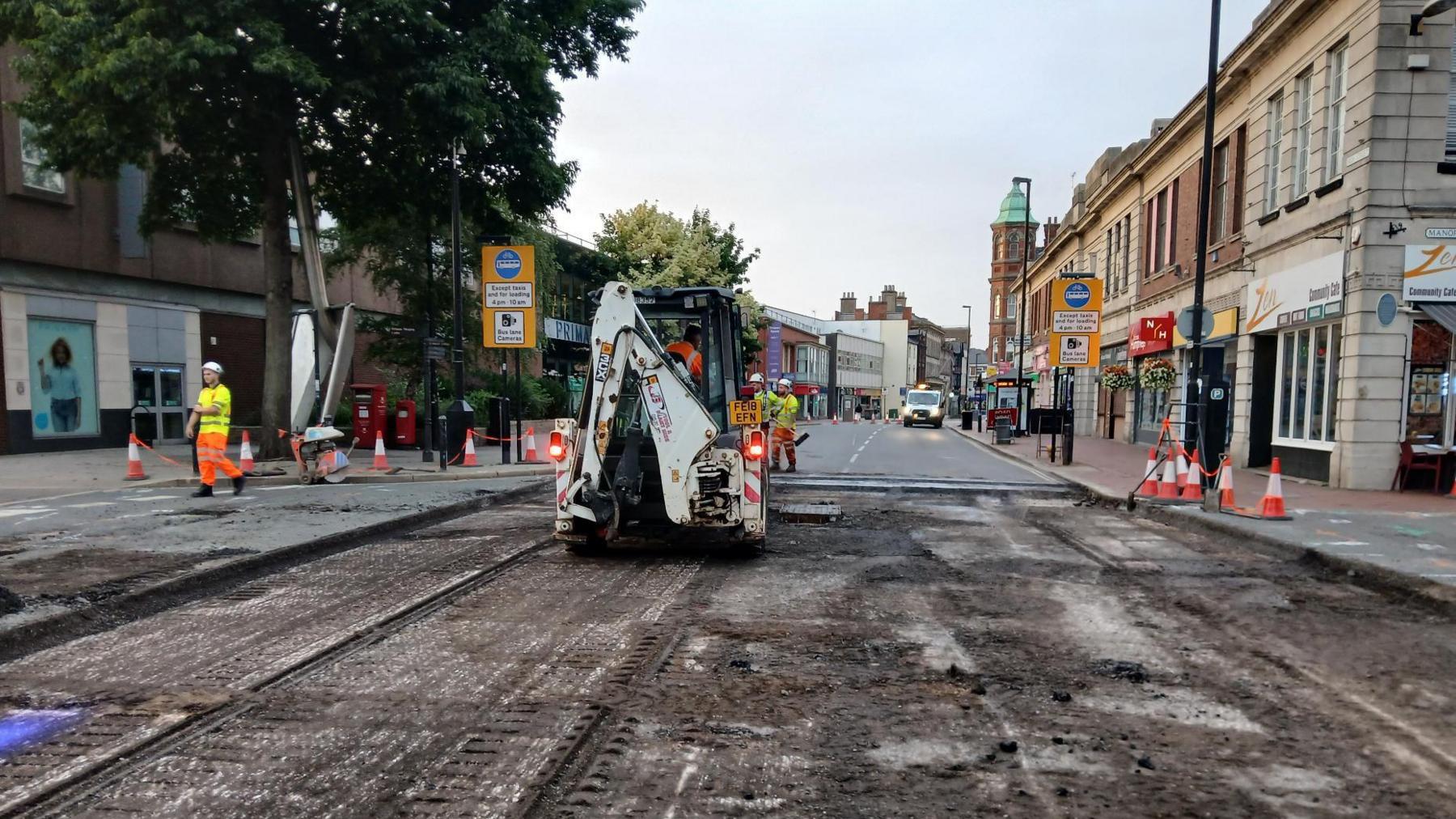 A town centre road which is having new tarmac put down. It is covered with mud and tracks. A white vehicle is being driven along it while workers in yellow and orange hi-vis outfits move about the site. High street buildings and trees are visible on either side of the road.