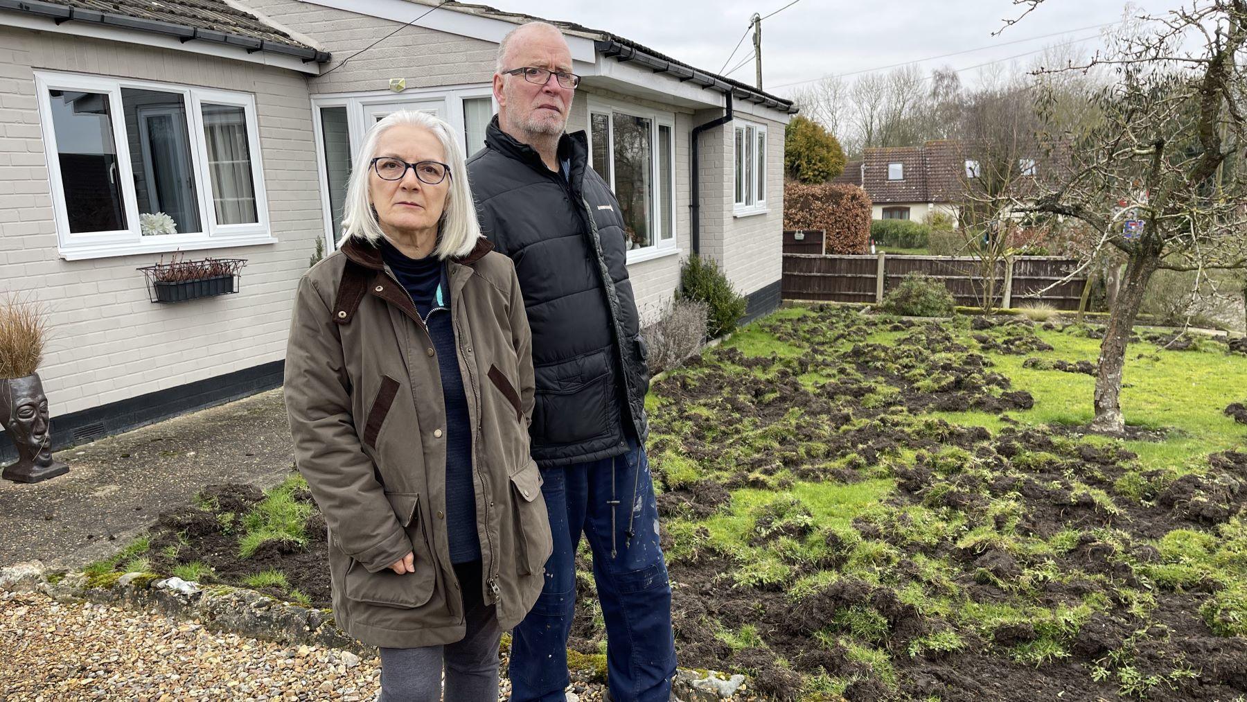 Lorraine and Phil Williams stood by their damaged front lawn. Lorraine, who has shoulder-length white hair and black glasses, is wearing a brown wax jacket. While Phil, who has short grey hair and thin glasses, is wearing a black puffer jacket and jeans.