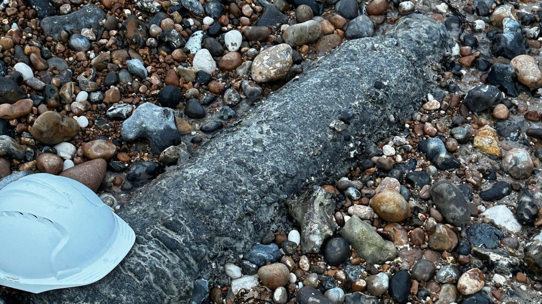 An unidentified long grey object in the shape of a large gas cannister. It is partially buried in stones on a beach. There is a white hard hat resting on the base of the object