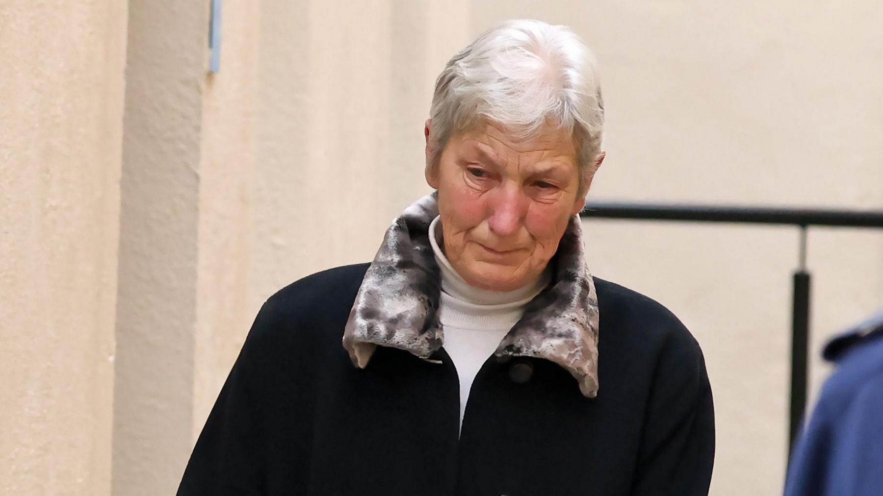 A woman with short white hair wearing a poloneck and high-collared jacket. She is leaning as though she is in the middle of take a step, and is looking away from the camera