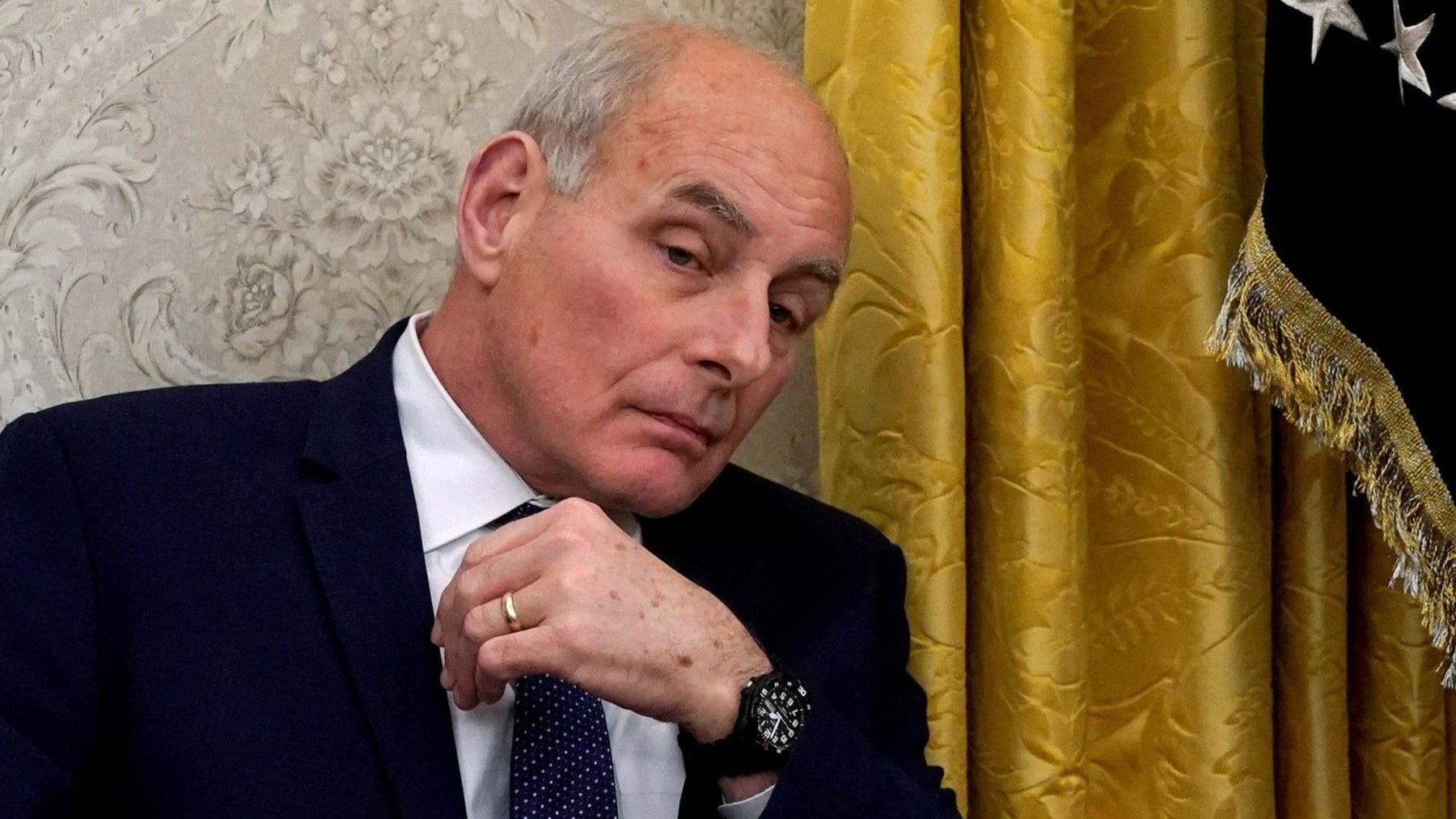 White House chief of staff John Kelly sits with a gold curtain in the background while wearing a blue suit and tie