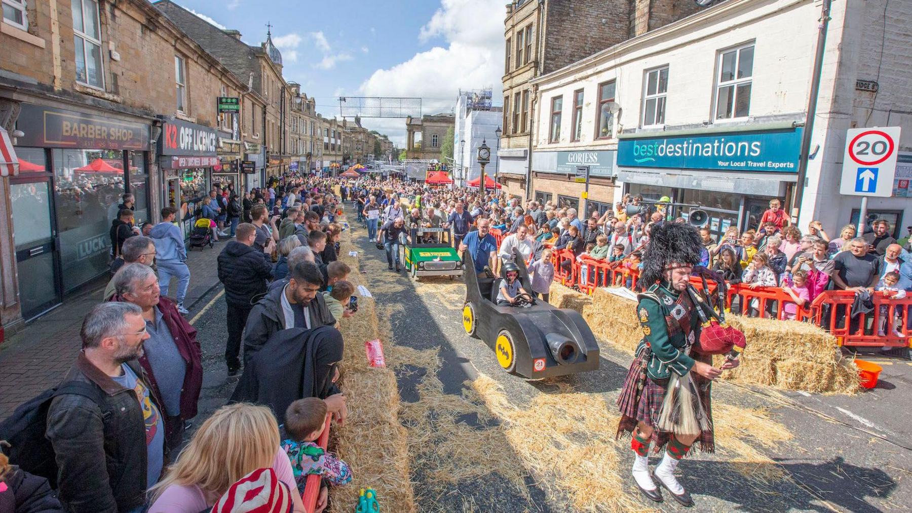 A piper leads the soapbox challenge contestants through Accrington town centre, including a Batman-themed soapbox. Hay bales are laid out to mark the race track, with crowds lining the streets to watch.