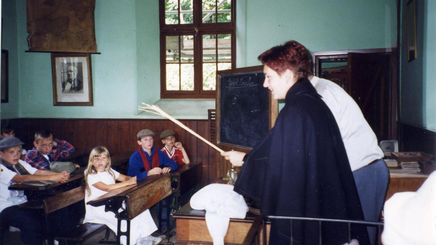 A woman stands at the front of an old classroom with a chalkboard behind her. Children, including Anton, sit at tables watching her.