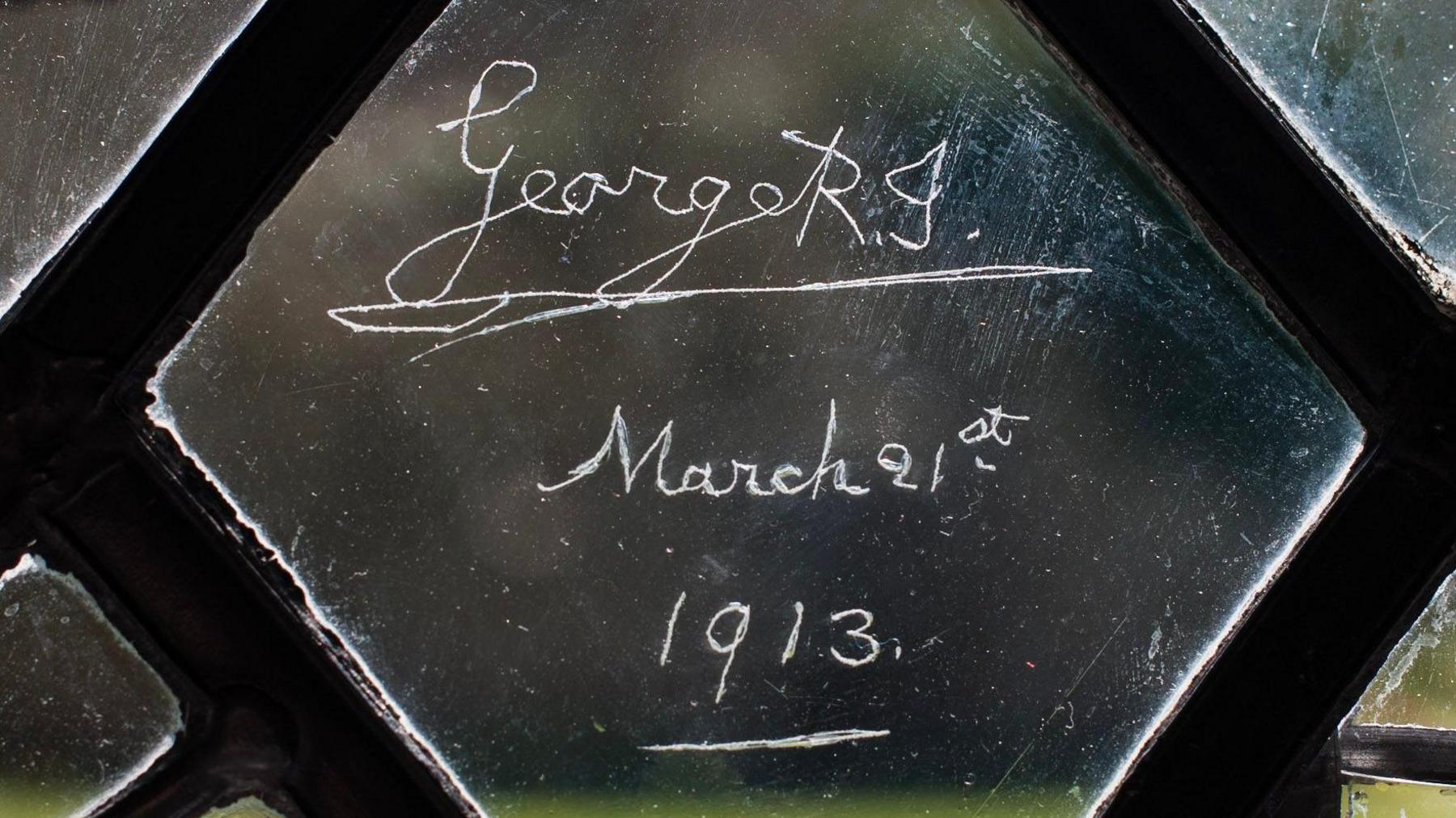 A small pane in a window. The pane is engraved with the signature of George V and the date March 21st 1913.